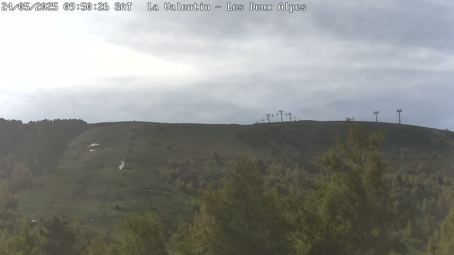 A mostly cloudy sky overlooks a grassy hillside with sparsely scattered trees and chairlifts in the distance.