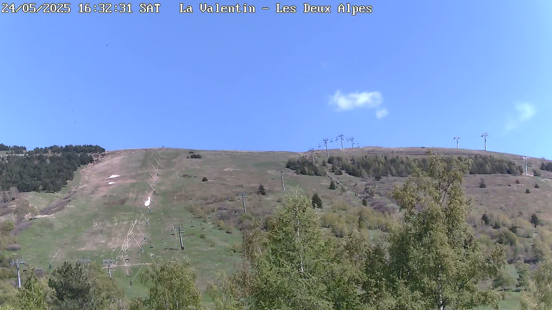 A mostly clear blue sky overlooks a grassy hillside dotted with trees and ski lifts, with a few patches of snow remaining.