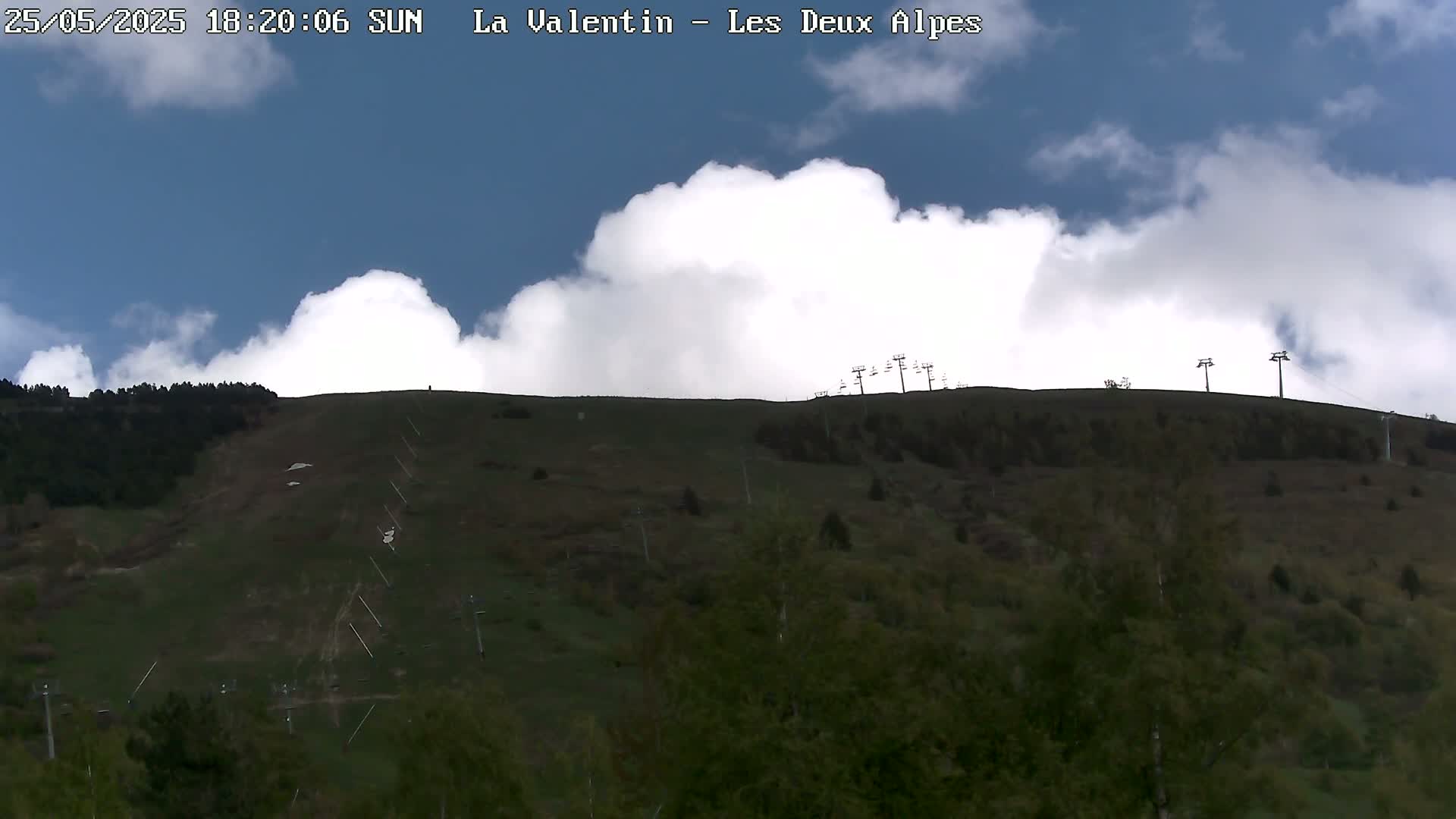 A partly cloudy sunny day shows a grassy hillside with ski lifts and sparse trees.