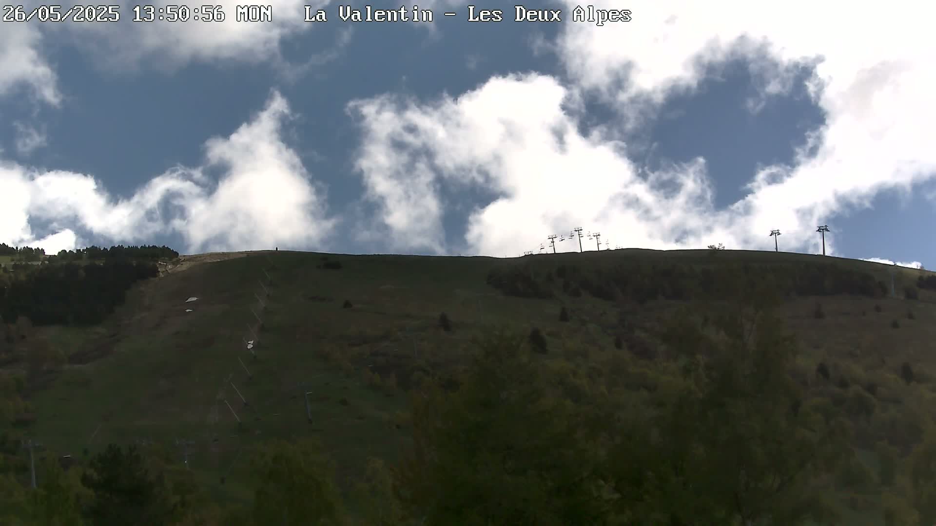 A partly cloudy sky overlooks a grassy hillside with sparse trees and a chairlift system.