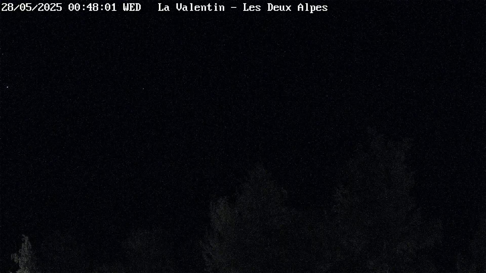 A nighttime, low-light image shows a dark, mostly featureless scene with some indistinct shapes possibly representing trees or vegetation in the lower portion.