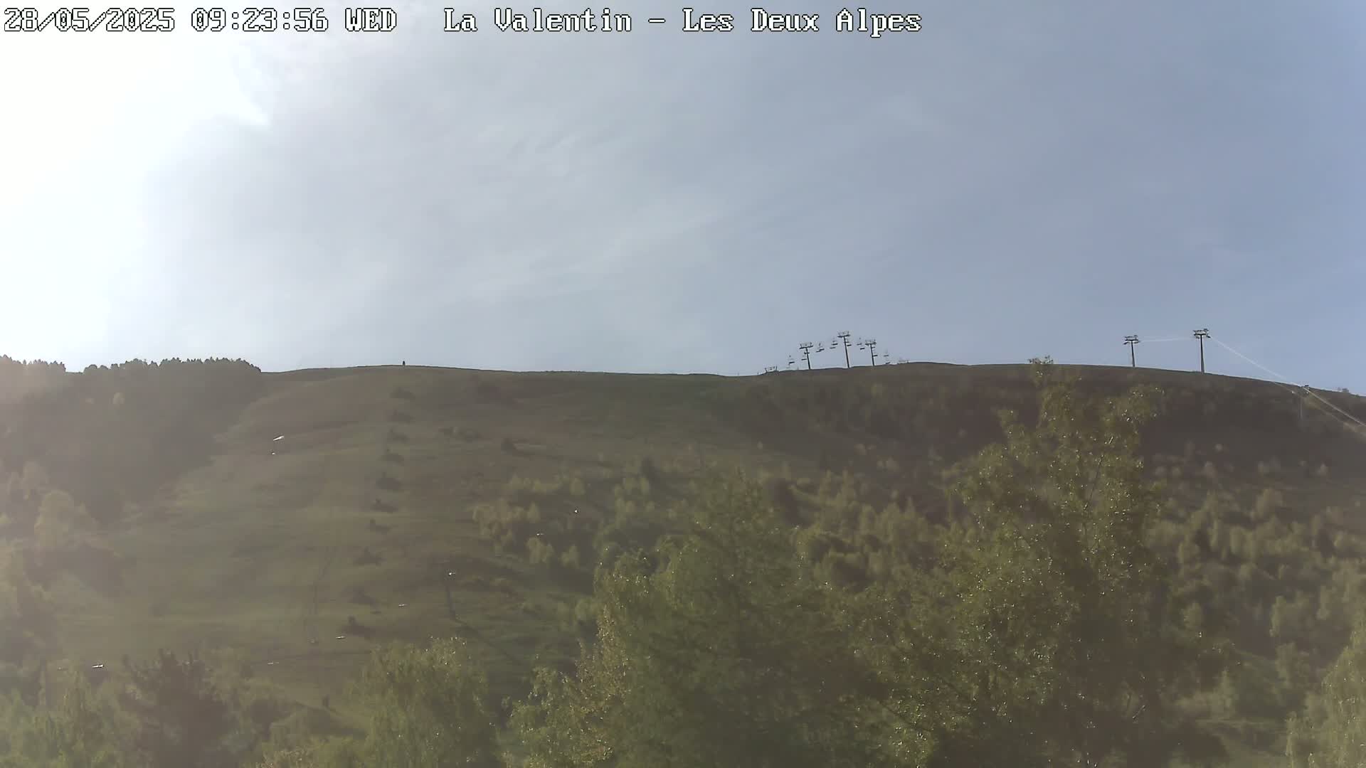 A mostly clear sky overlooks a grassy hillside with sparse trees and a chairlift at the top.