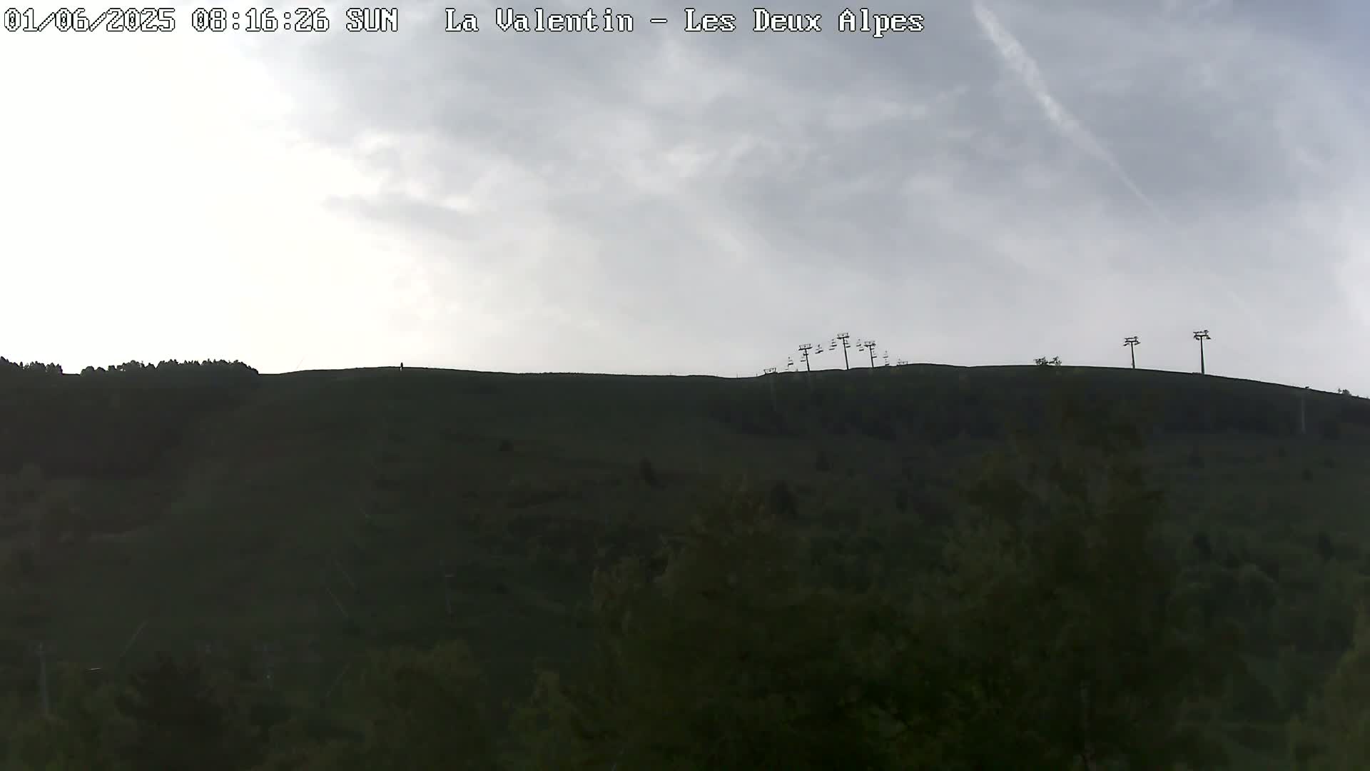 A mostly cloudy sky overlooks a dark green hill with several chairlift towers on its crest.