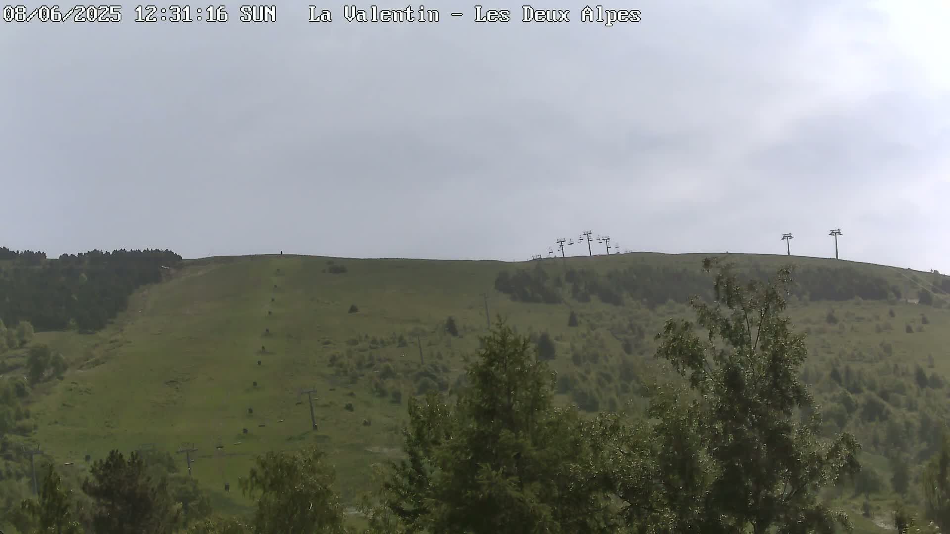 A mostly cloudy day reveals a grassy hillside with sparse trees and chairlifts atop it.