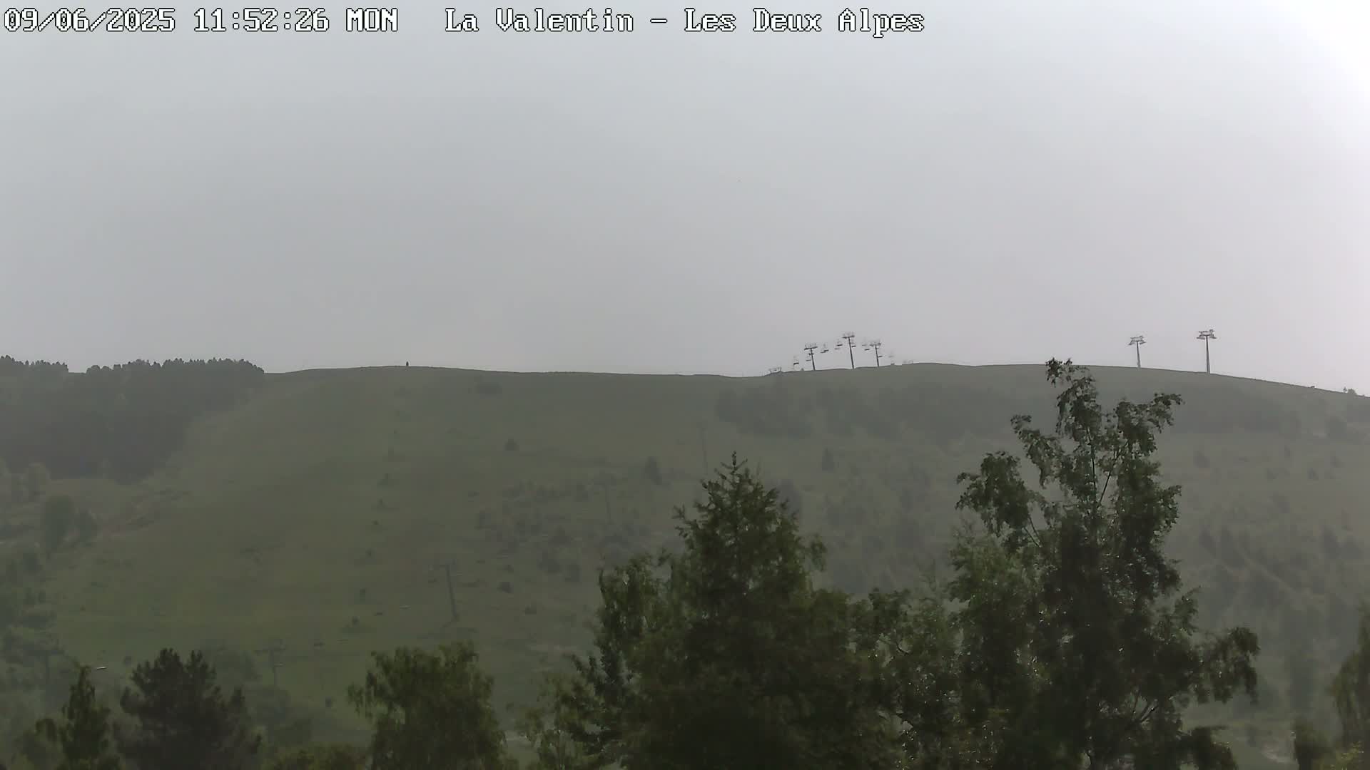 A hazy, overcast view shows a green hill with several chairlift towers and some trees in the foreground.