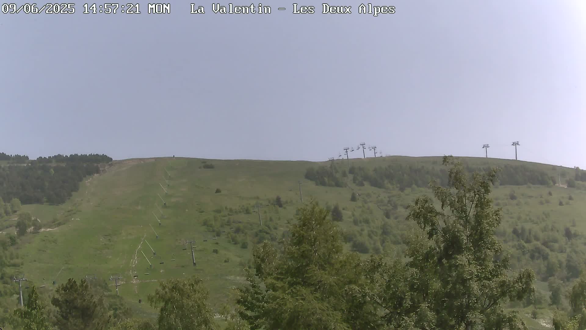 A grassy hillside with several chairlifts and some trees under a hazy sky.