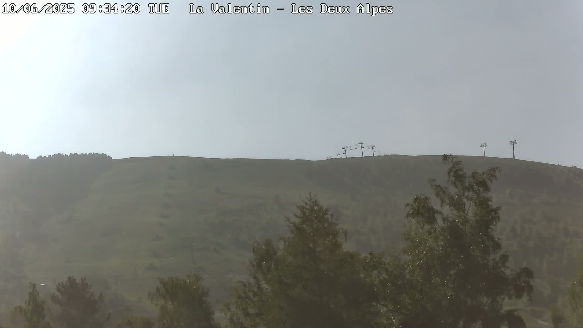 A hazy view of a grassy hill with chairlifts at the top, partially obscured by trees in the foreground.