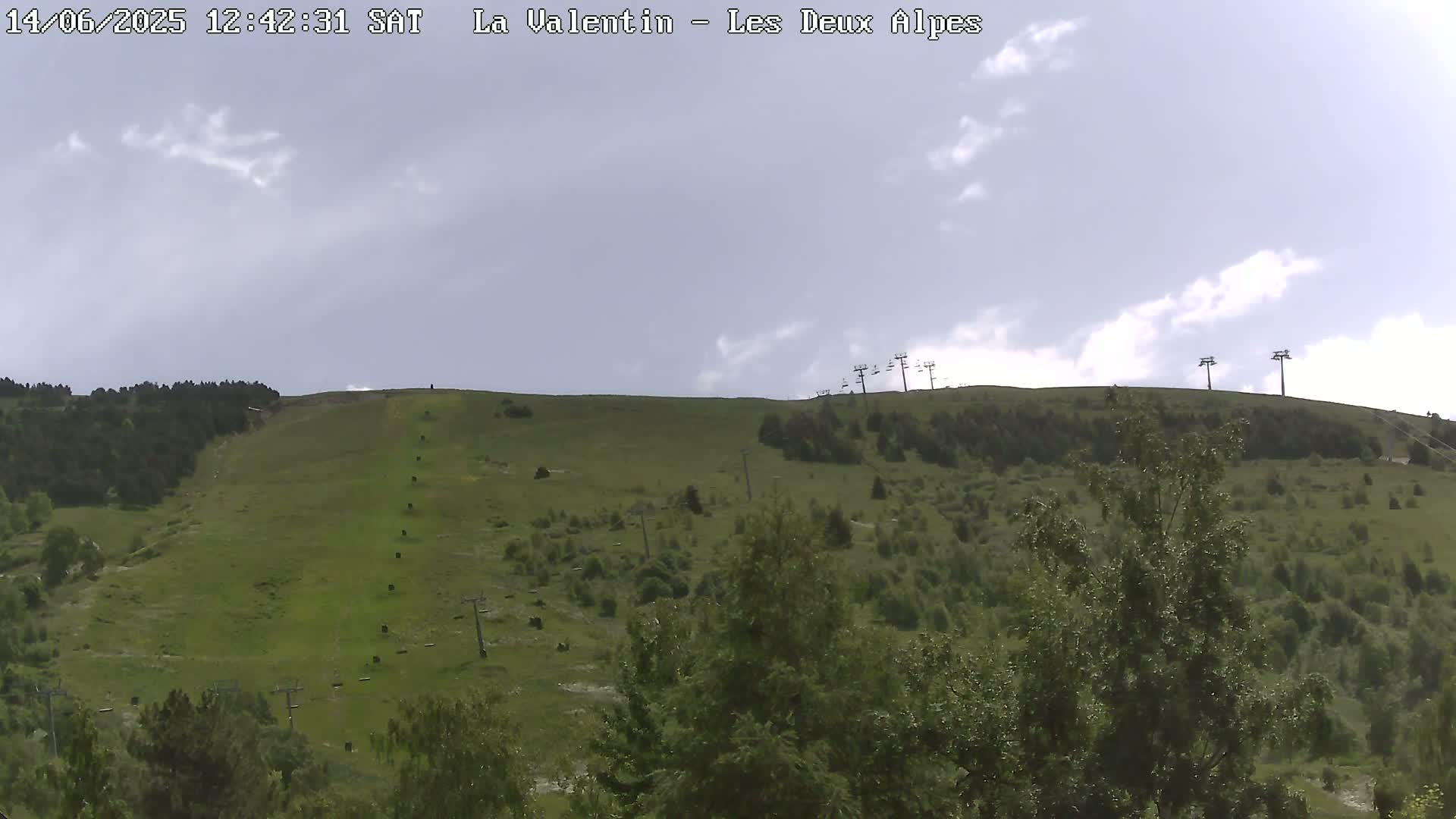 A mostly cloudy sky overlooks a grassy hillside with several ski lifts and scattered trees.