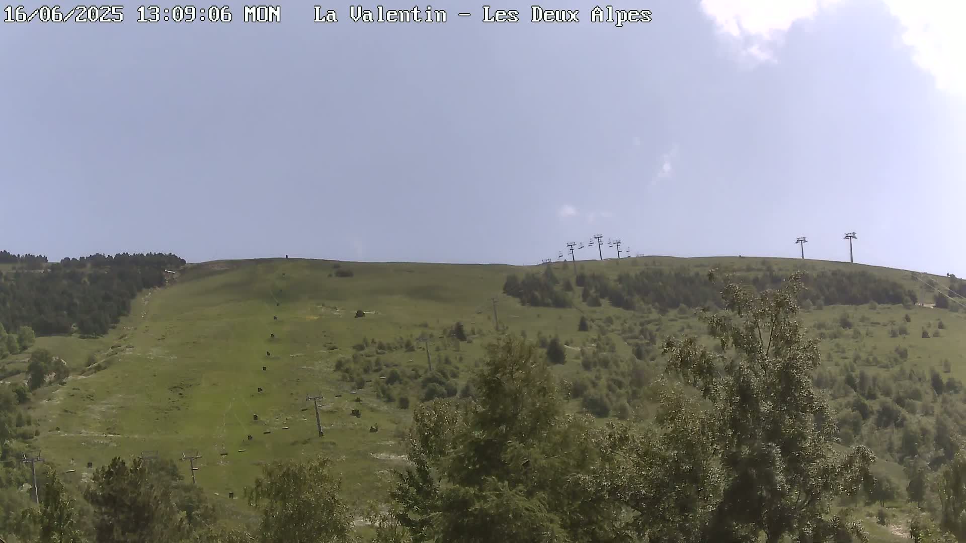A mostly sunny day reveals a grassy hillside dotted with trees and several chairlifts, some of which appear to be inactive.