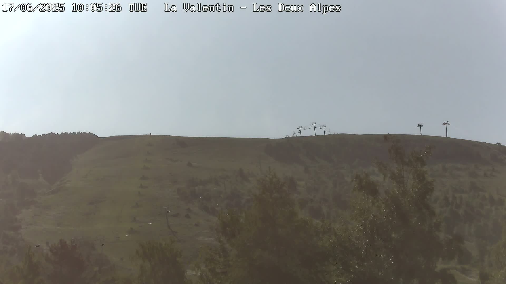 A hazy view shows a grassy hill with a few sparse trees and chairlifts at the top.