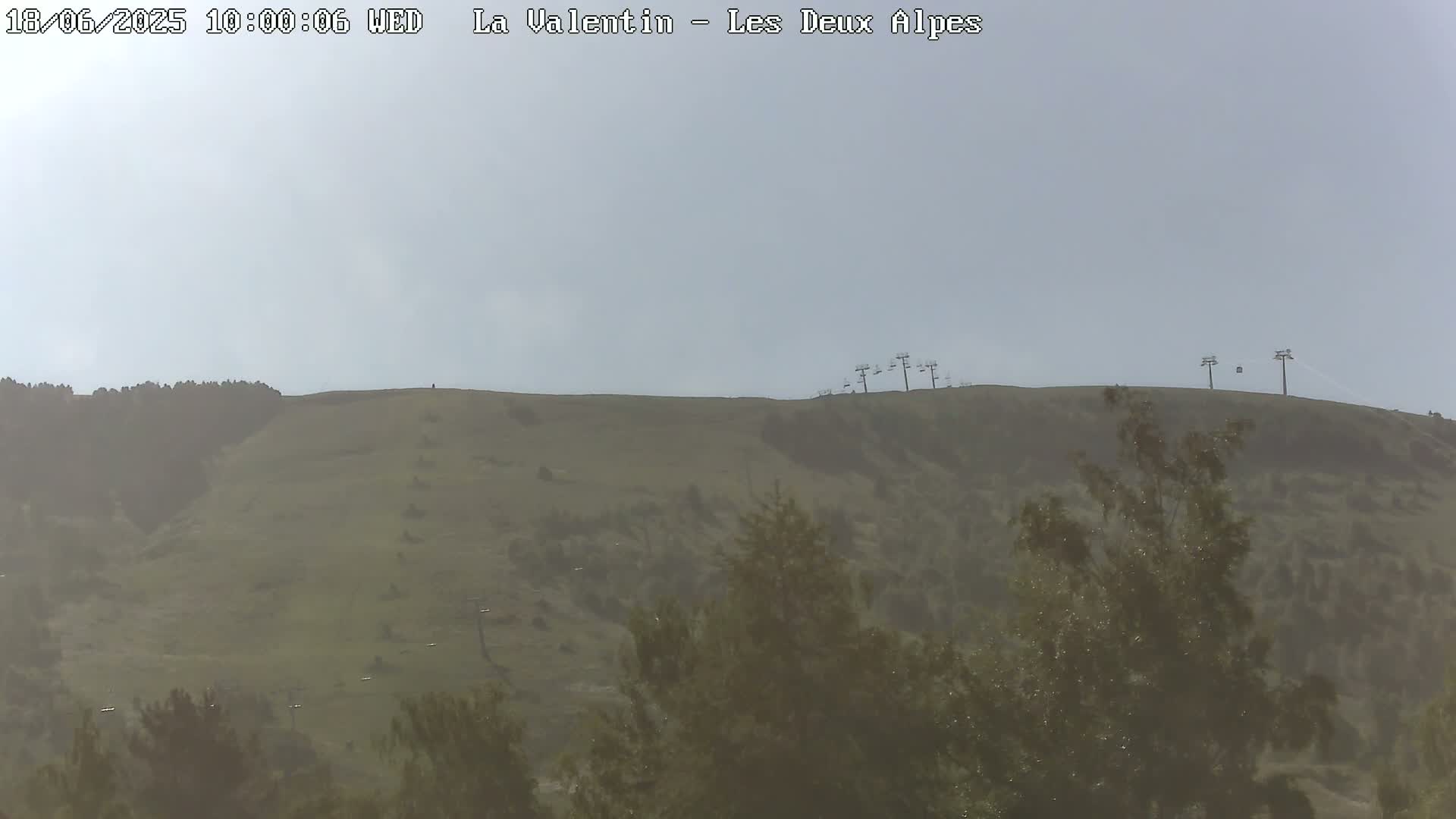 A hazy view of a grassy hill with several ski lifts atop it and trees in the foreground.