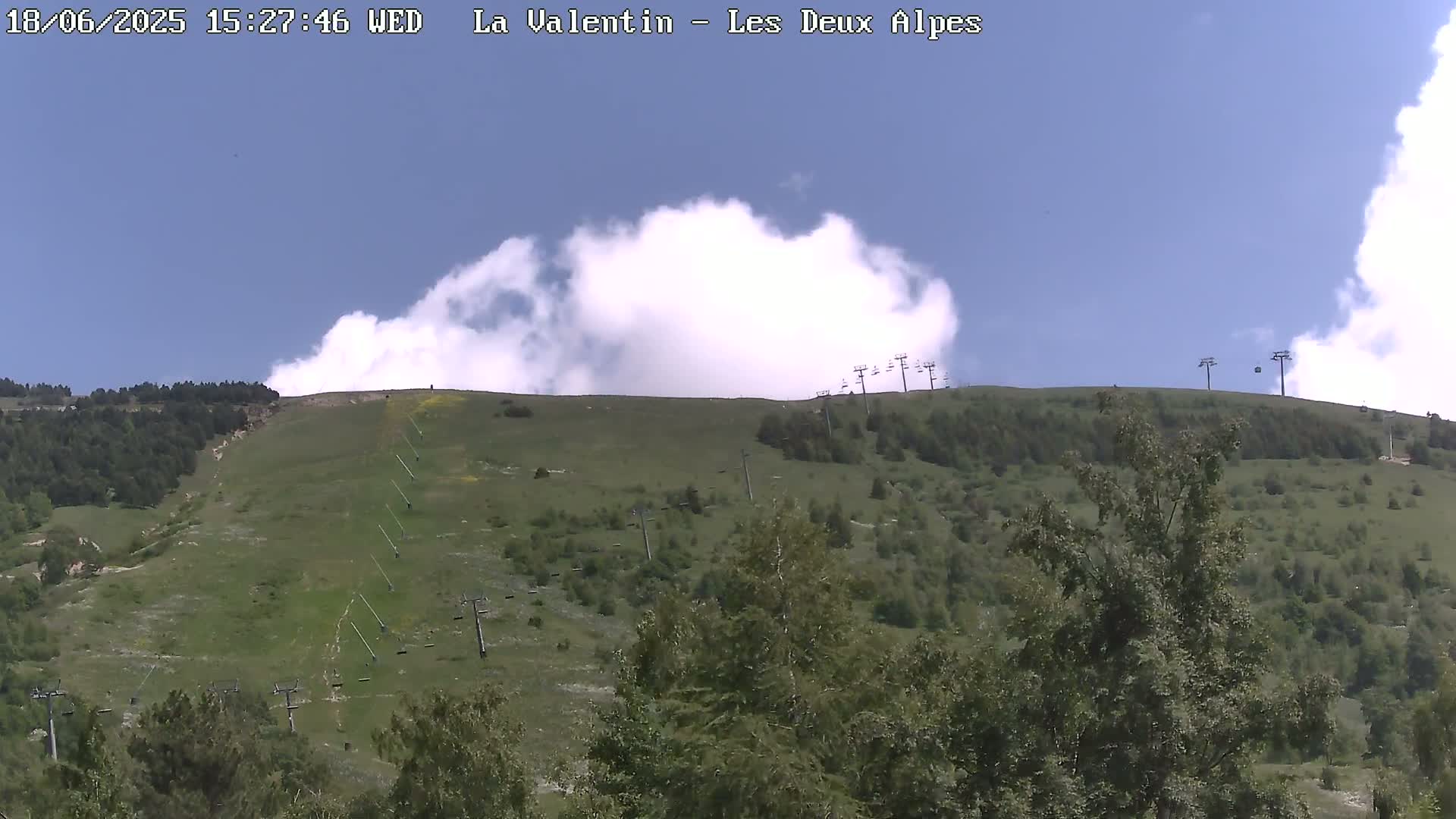 A partly cloudy sky overlooks a grassy hillside dotted with trees and several chairlifts.