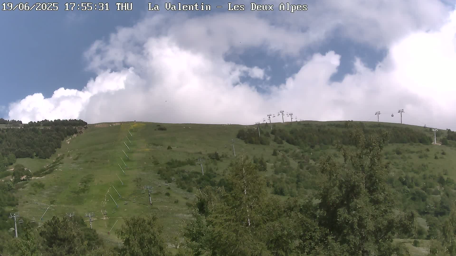 A partly cloudy sky overlooks a grassy hillside with several chairlifts and scattered trees.