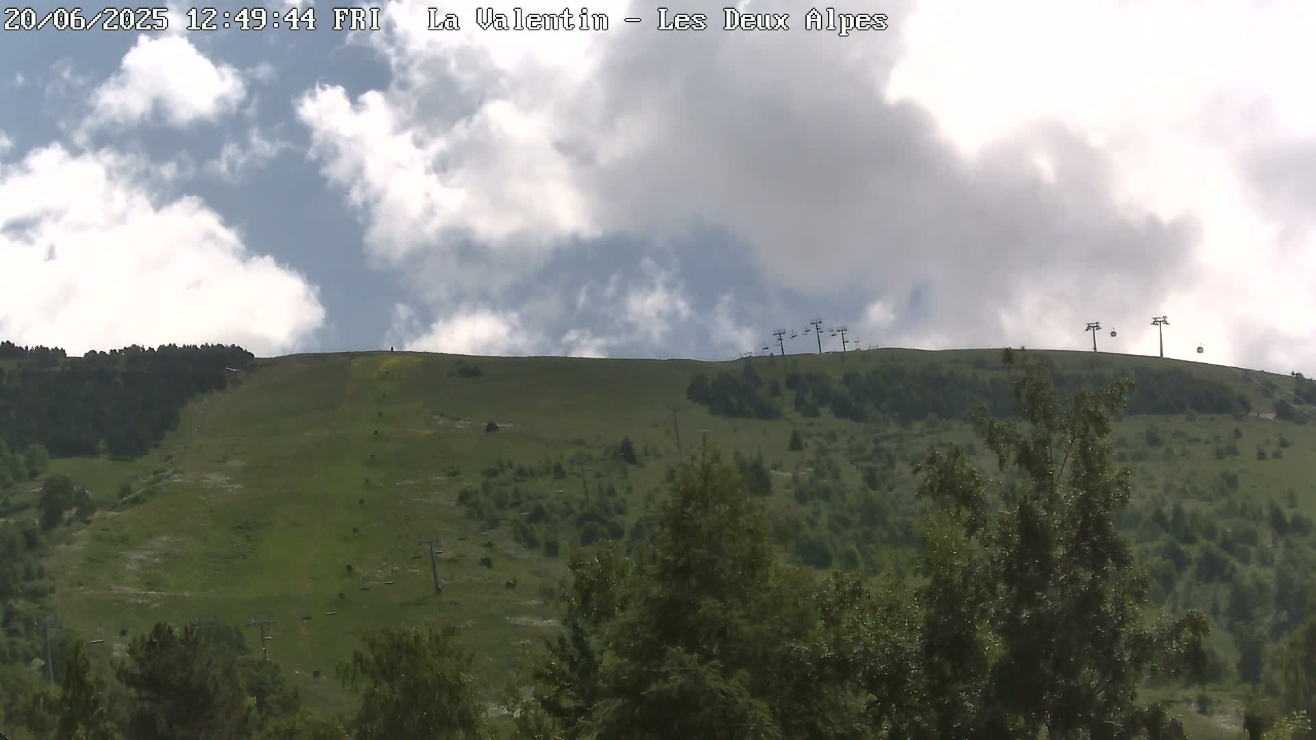 A partly cloudy sky overlooks a grassy hillside with scattered trees and several chairlifts.