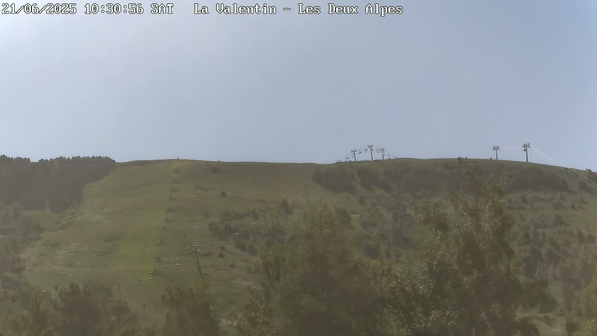 A hazy, mostly clear day reveals a grassy hillside with a few trees and chairlifts in the distance.
