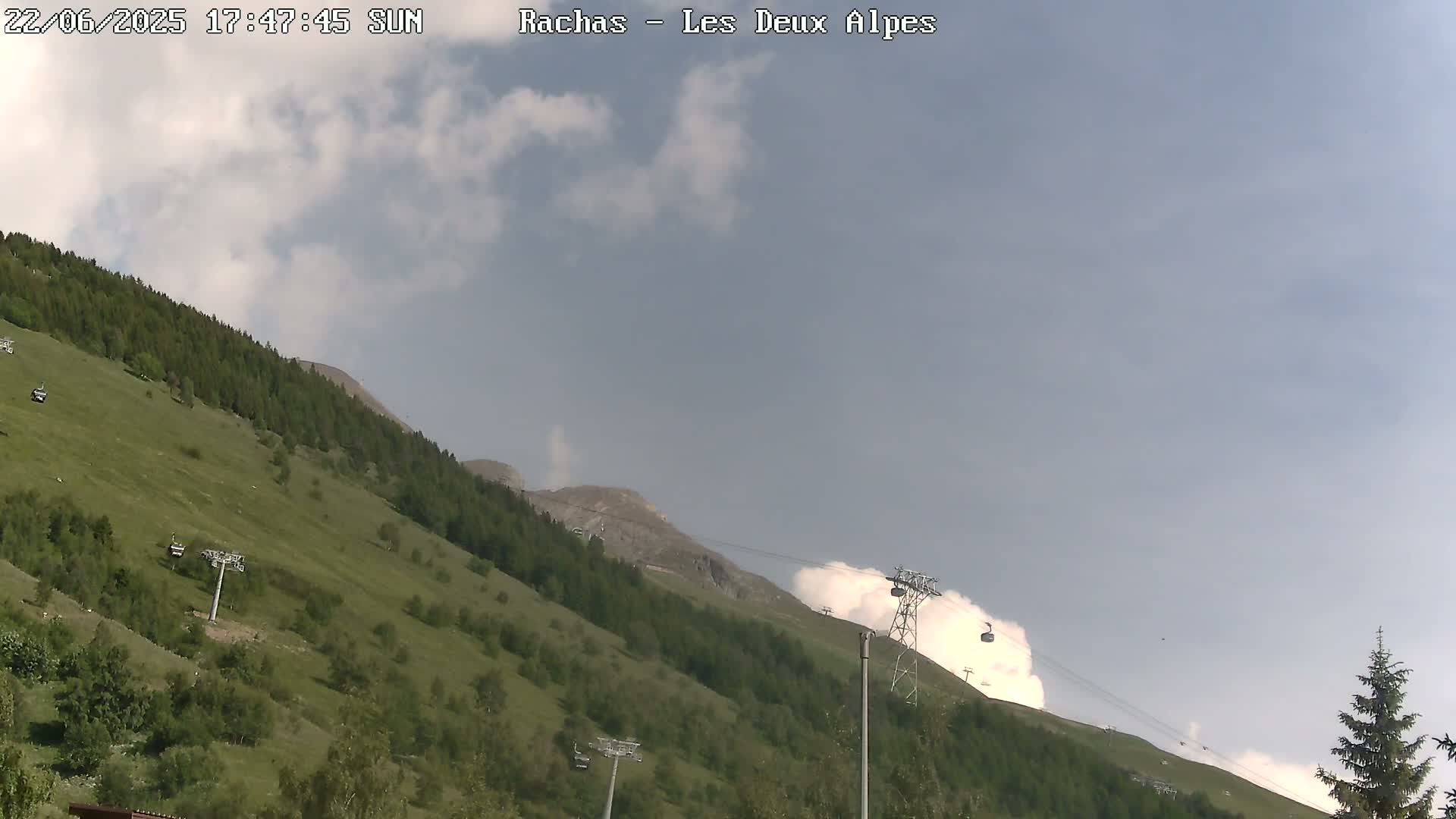 A mostly sunny, partly cloudy day shows a grassy mountainside with chairlifts and a mountain peak in the background.