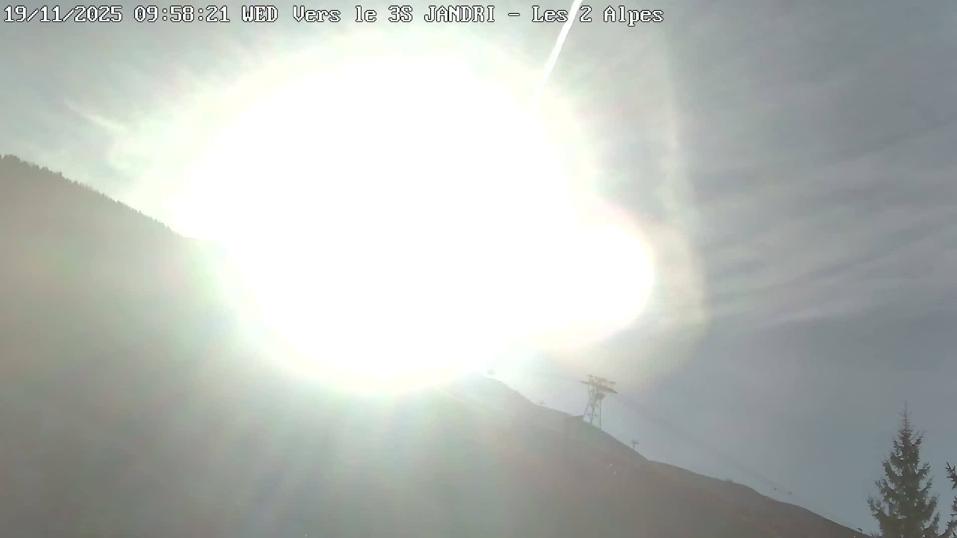 A bright sun causes significant glare over a mountain landscape featuring a distant ski lift tower and a coniferous tree under a partly hazy sky.