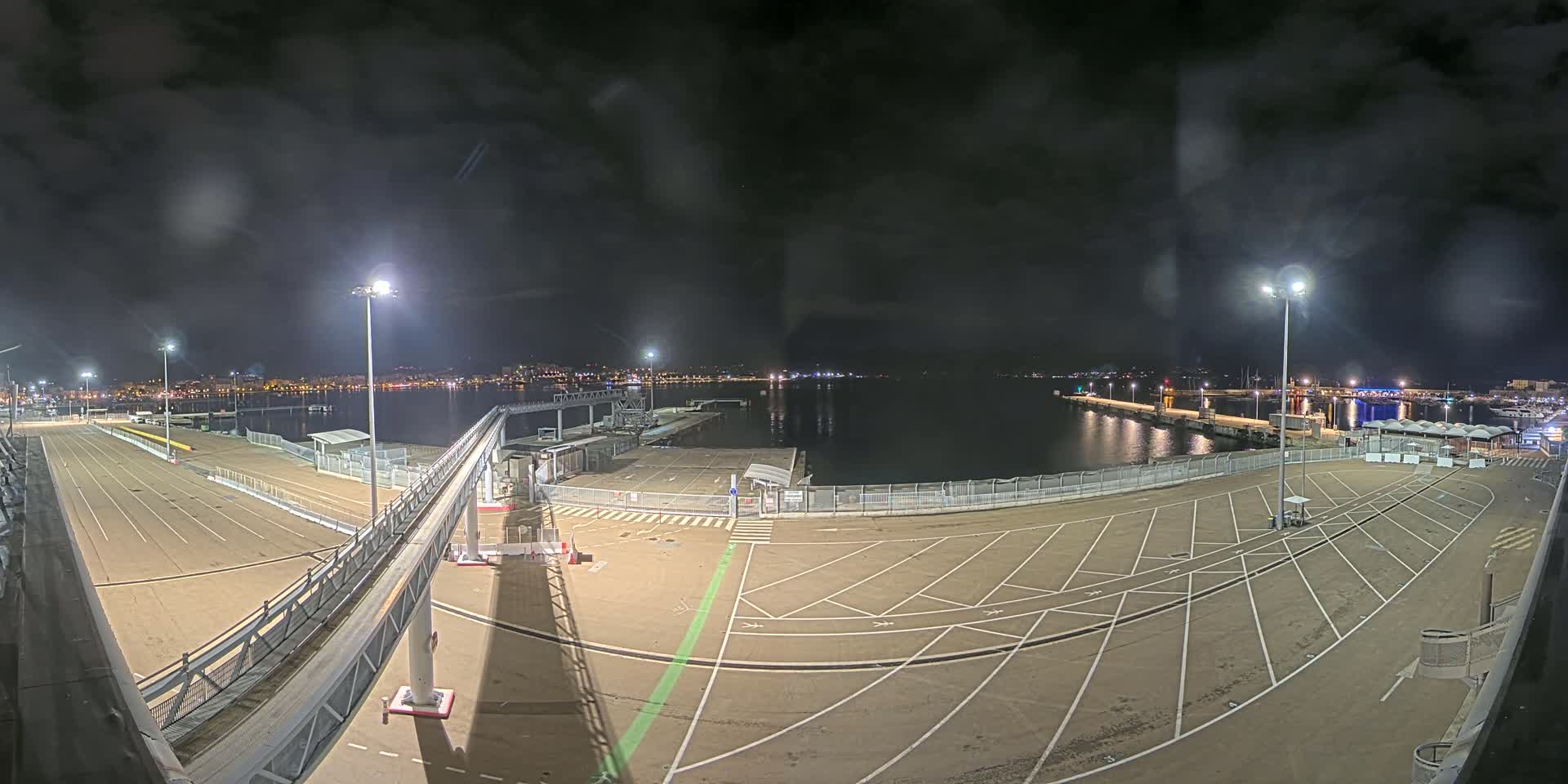 A nighttime view of an empty ferry terminal parking lot with a gangway leading to a dock, overlooking a calm body of water and a city skyline in the distance under a cloudy sky.