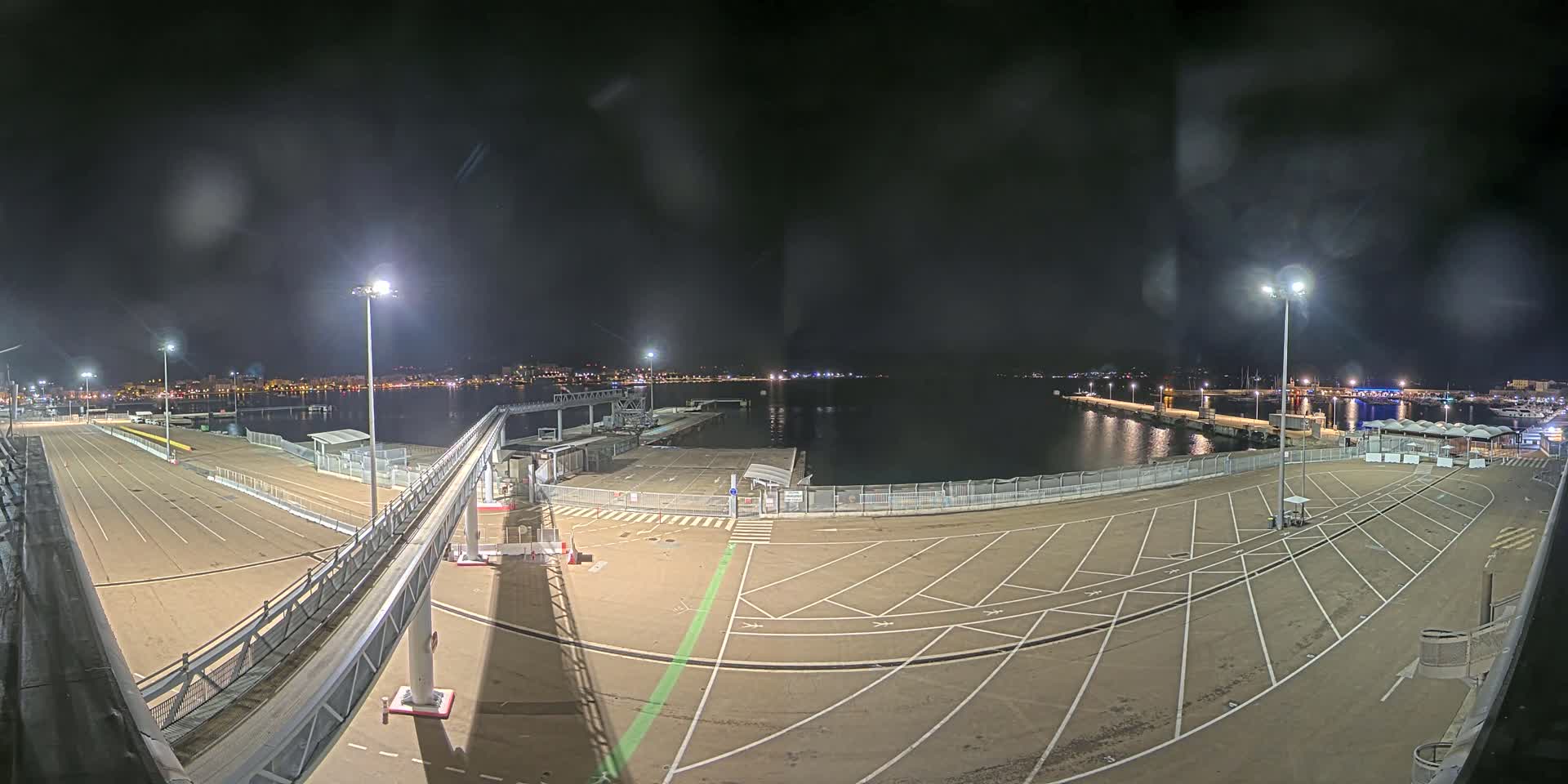 An empty, nighttime port area with a passenger gangway, lined with parking spaces and illuminated by several tall lights, overlooks a calm body of water with a city skyline visible in the distance.