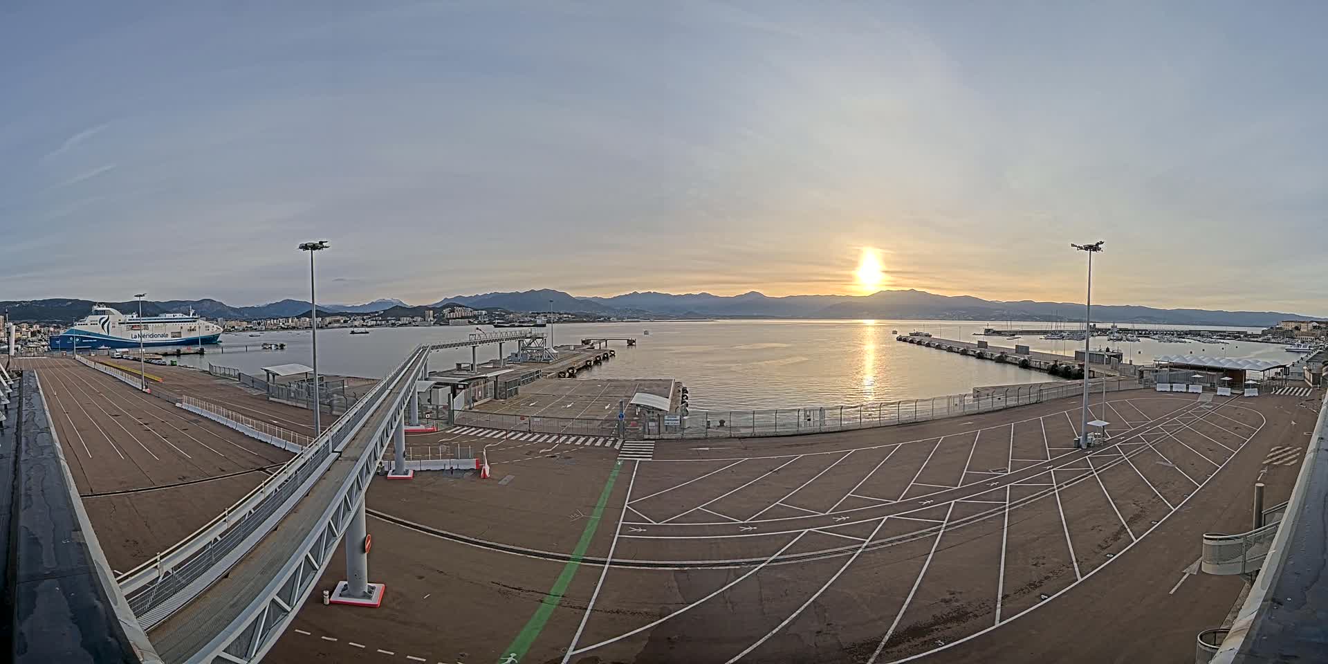 A ferry is docked at a port with a large empty parking lot under a clear sunset sky, mountains visible in the distance.