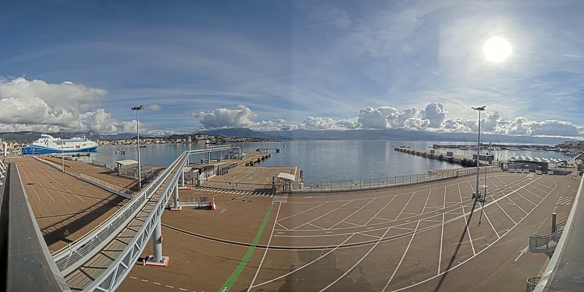 A ferry is docked at a port under a partly cloudy, sunny sky, with a large empty parking lot and walkways visible.