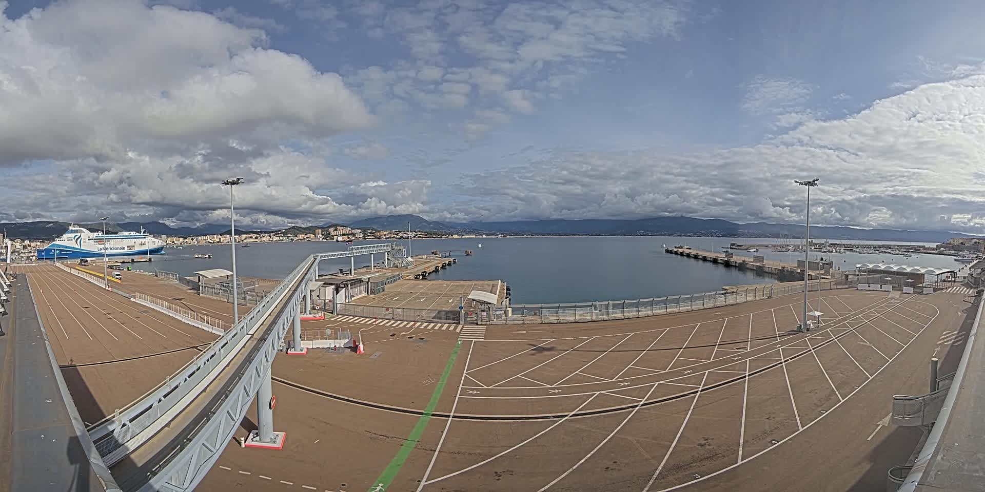 A ferry is docked at a port under a partly cloudy sky, with a large empty parking lot and a walkway leading to the ship.