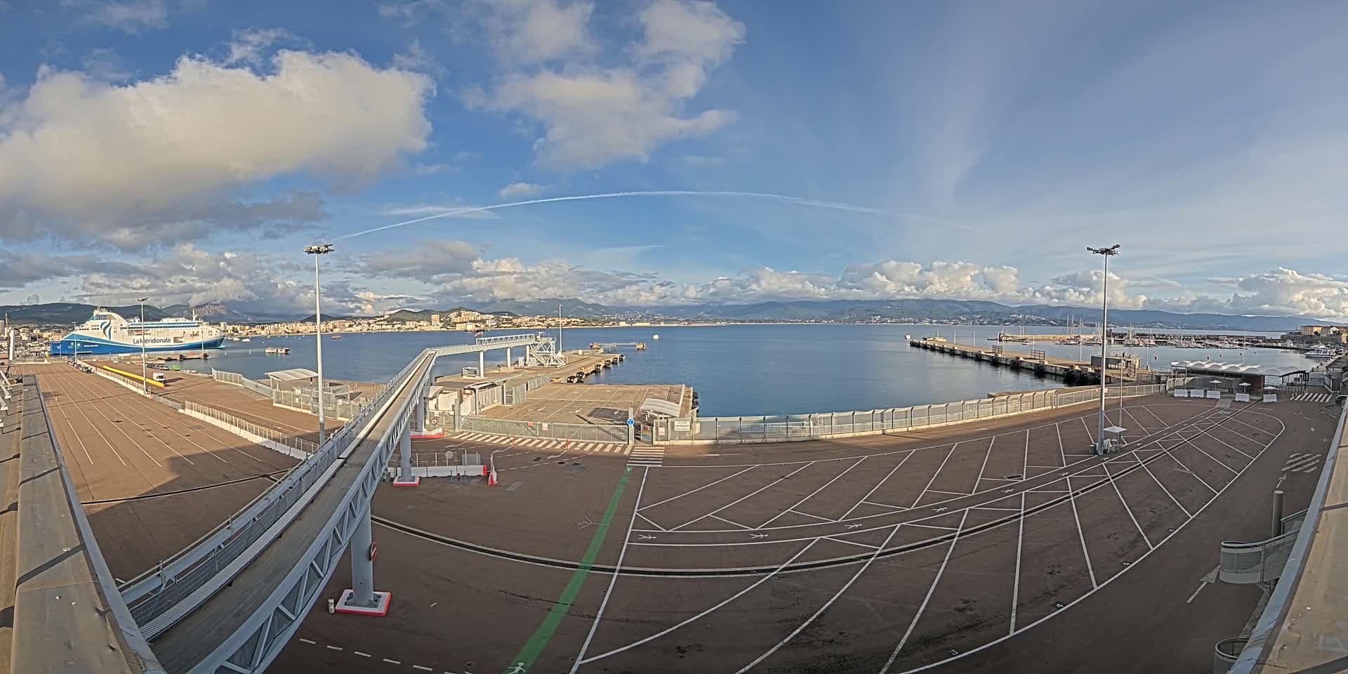 A ferry is docked at a port under a partly cloudy sky, with a large empty parking lot and a walkway leading to the ship.