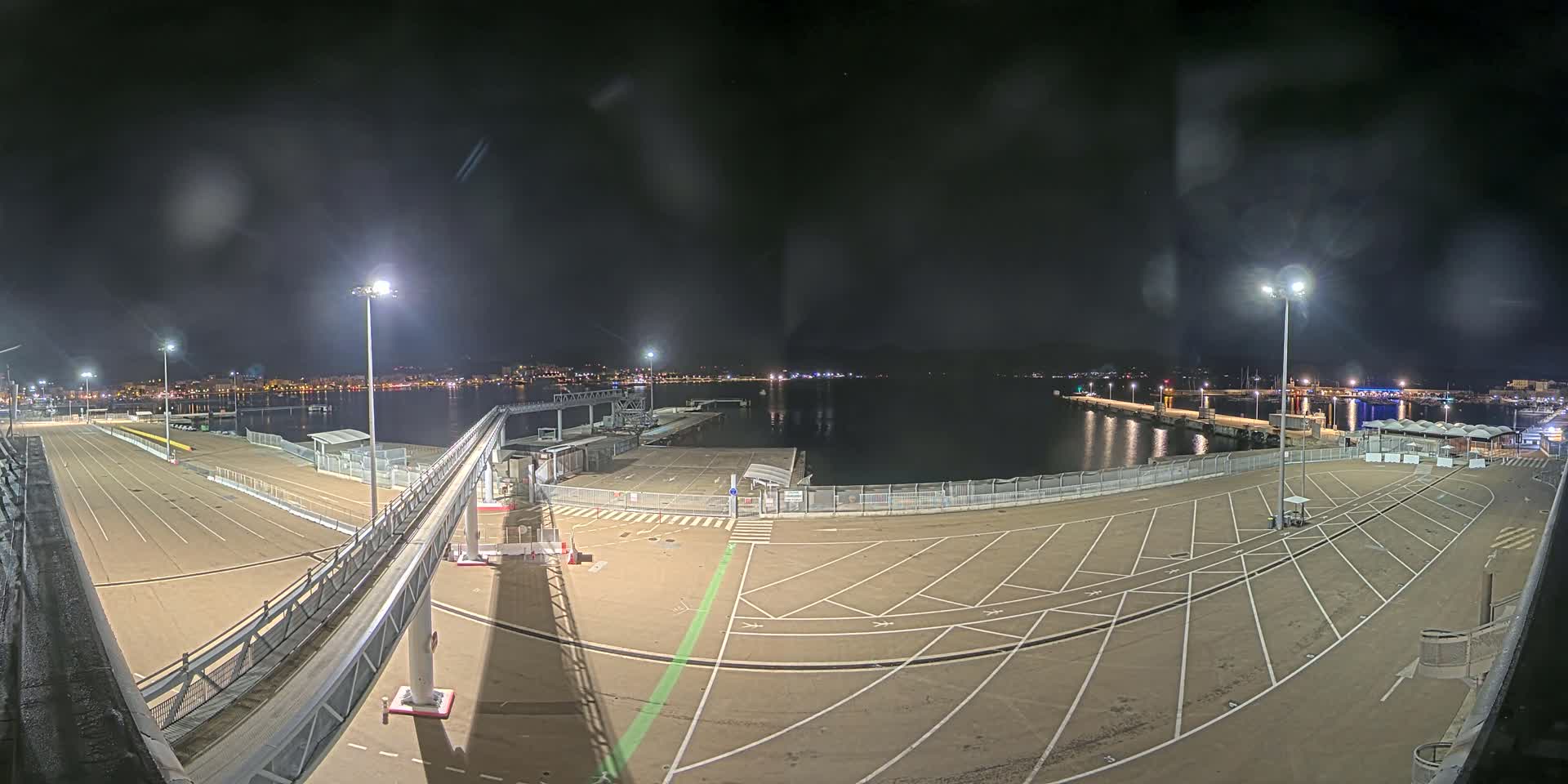 A nighttime view of an empty port area with a walkway and parking spaces, overlooking calm water and a city skyline in the distance.