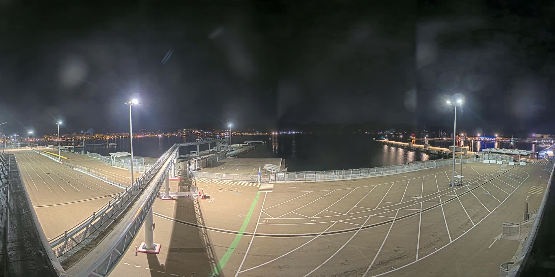 An empty nighttime port parking lot with a gangway leading to a dock, illuminated by bright lights, overlooks a calm body of water and a distant city skyline.