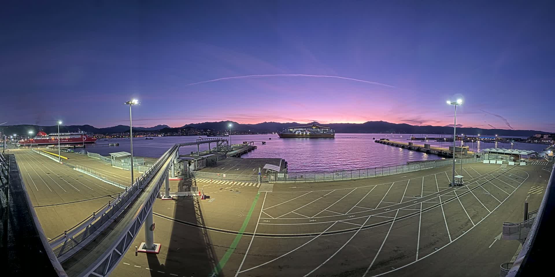A large empty parking lot at a ferry terminal overlooks a calm harbor with two ferries docked, all under a twilight sky with purple and pink hues.