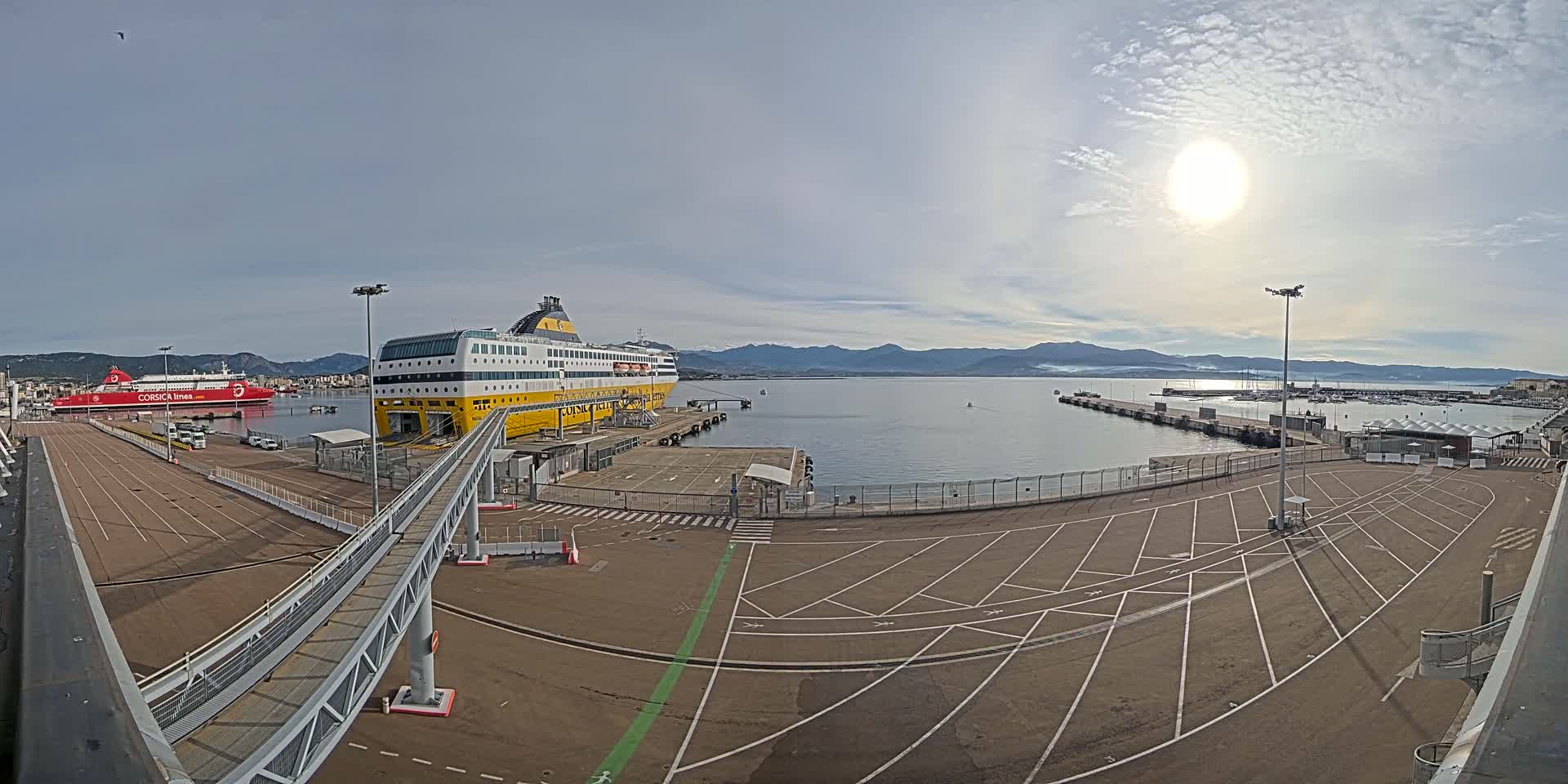 A large yellow and white ferry is docked at a port on a sunny day with calm water and mountains in the distance.
