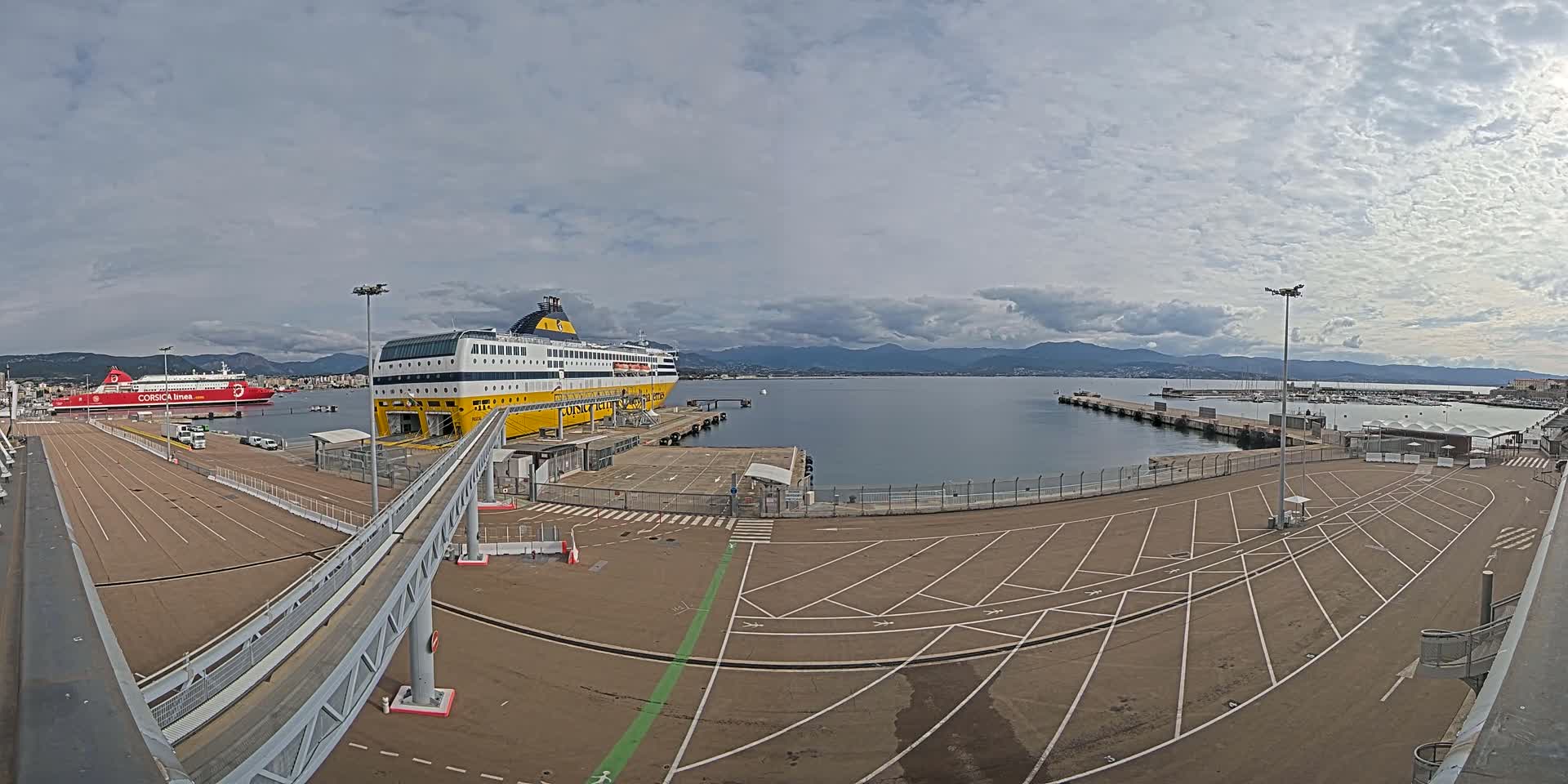A large yellow and white ferry is docked at a port under a cloudy sky, with a smaller red ferry visible in the background.
