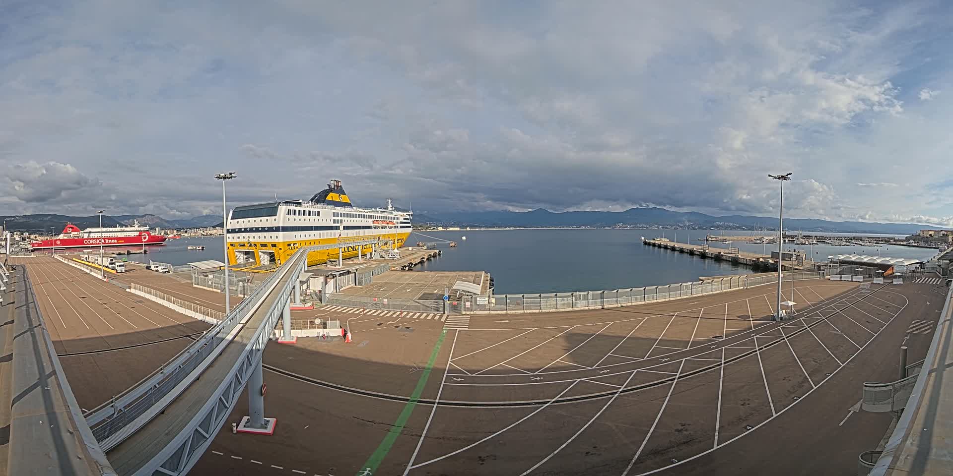 A large yellow ferry is docked at a port under a partly cloudy sky, with another smaller red ferry visible in the background.