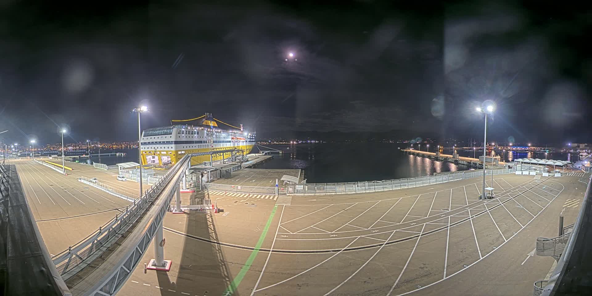 A large ferry is docked at a port at night under a partly cloudy sky with a bright moon.