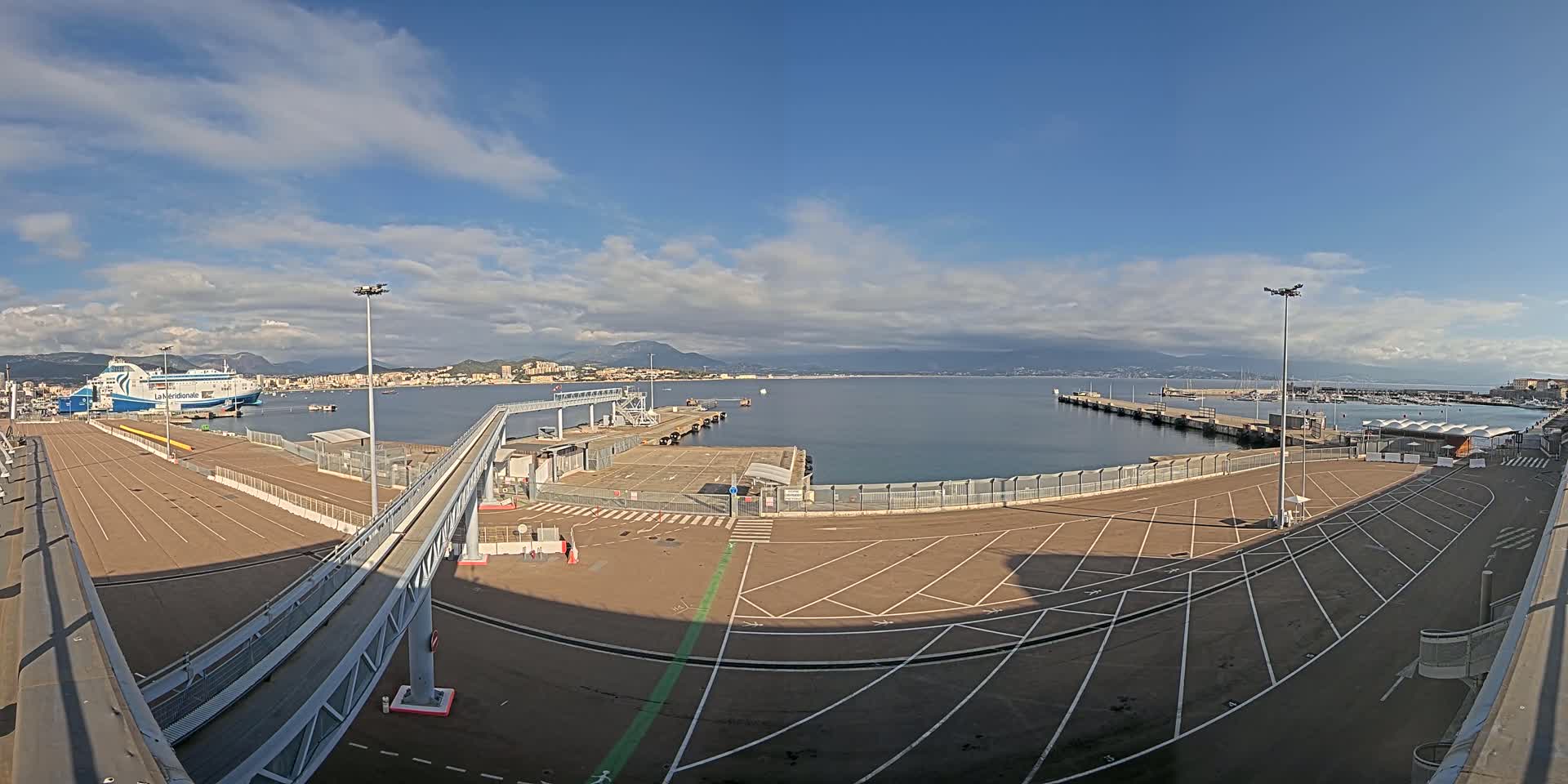 A ferry is docked at a port with a mostly sunny sky and a calm sea, and there's a large empty parking lot near a pedestrian walkway leading to the ship.