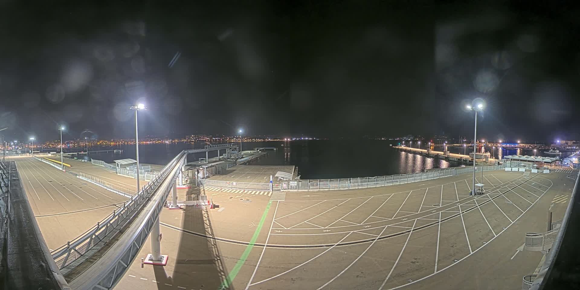 A nighttime view of an empty port area with a long walkway leading to a dock, illuminated by several tall lights, overlooking a calm body of water with a city skyline in the distance.