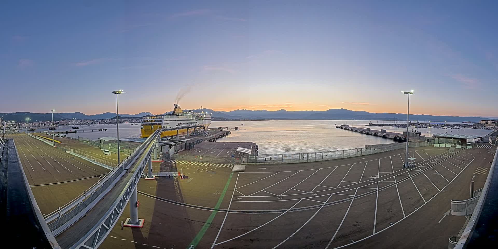 A large ferry is docked at a port at sunrise, with a walkway leading to it and a mostly empty parking lot visible.