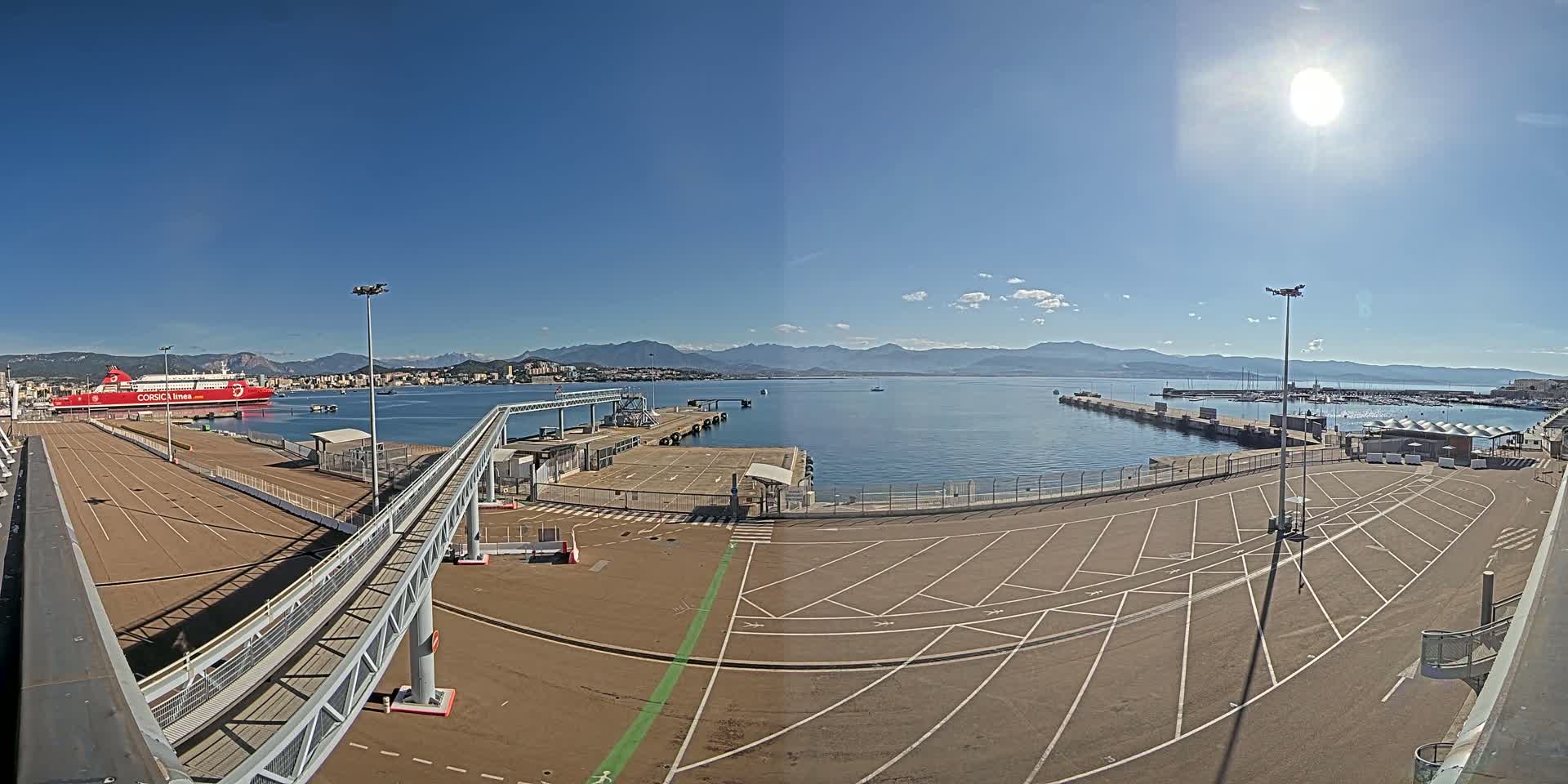 A large red ferry is docked at a port on a sunny day, with mountains visible in the distance across calm blue water.