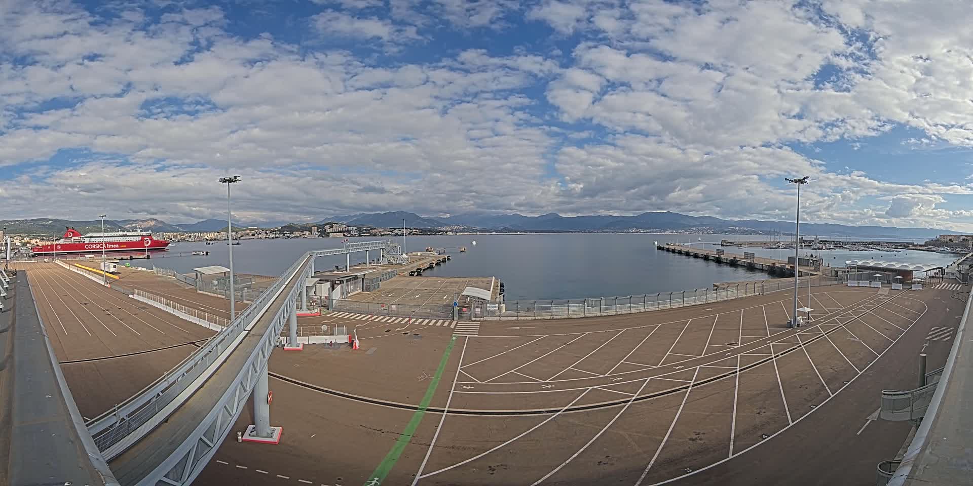 A large ferry is docked at a port under a partly cloudy sky, with mountains visible in the distance.