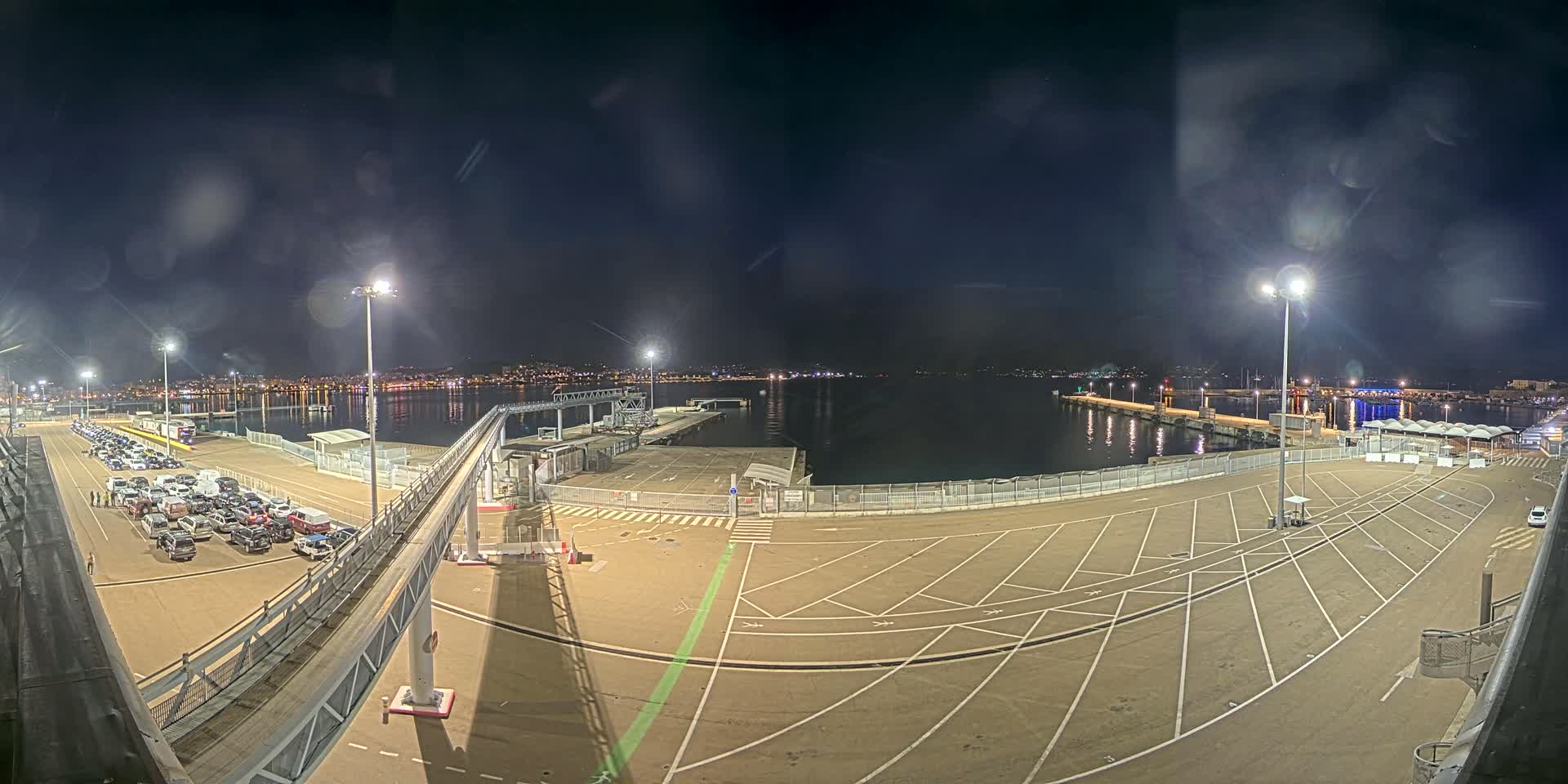 A nighttime view of a port with a large empty parking lot, a gangway, and numerous cars parked nearby, overlooking a calm body of water and a distant city skyline.