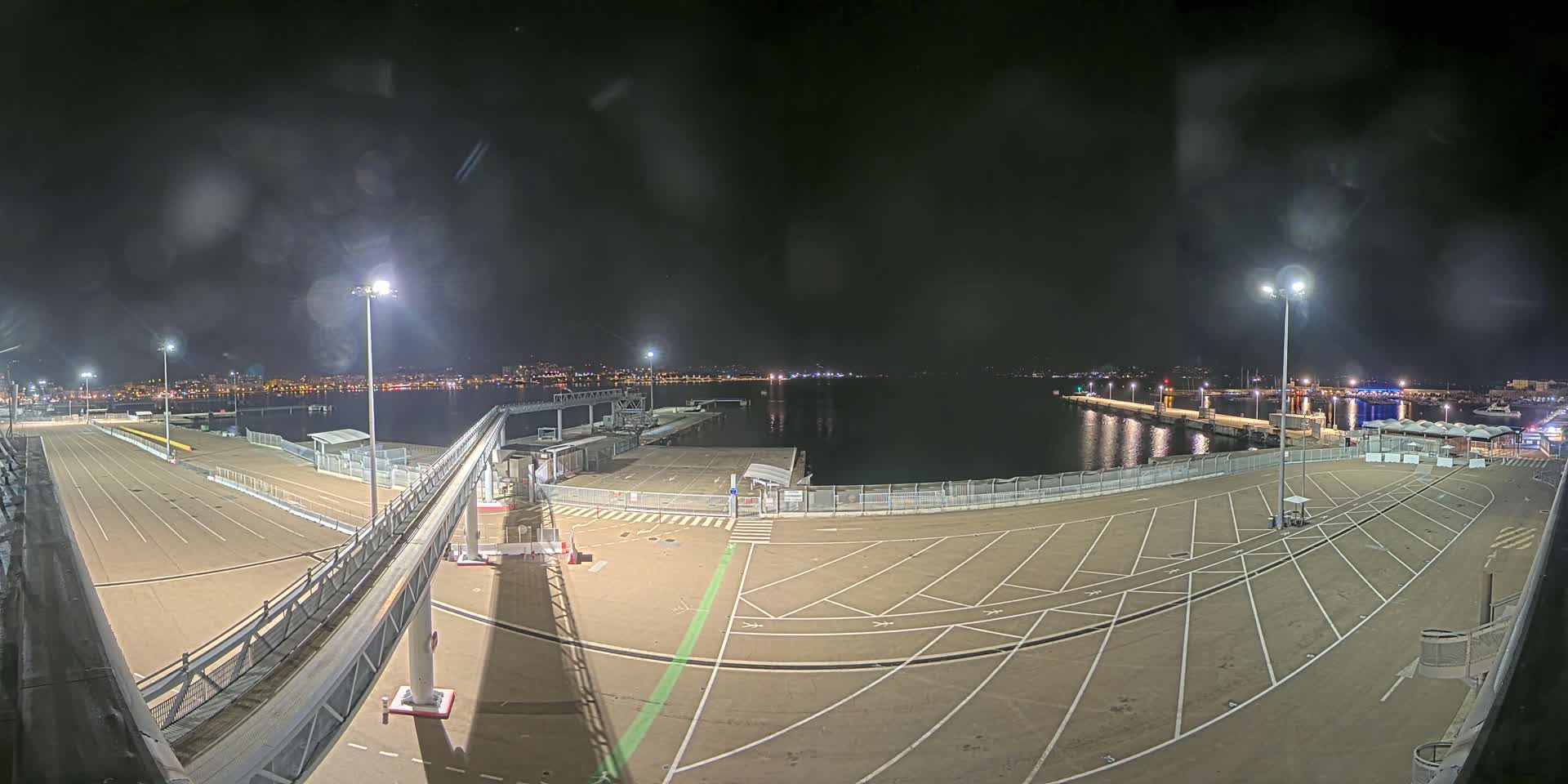A nighttime view of a mostly empty port area with a gangway and parking lot, illuminated by lights, overlooking calm water and a distant city skyline.