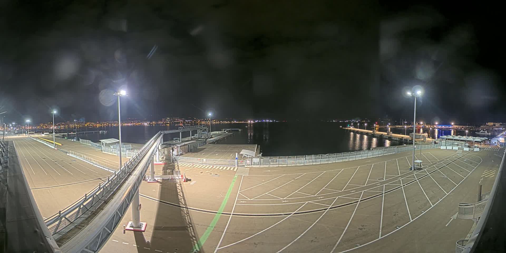 A nighttime view of a mostly empty port with a gangway leading to a dock, under slightly overcast skies and rain.