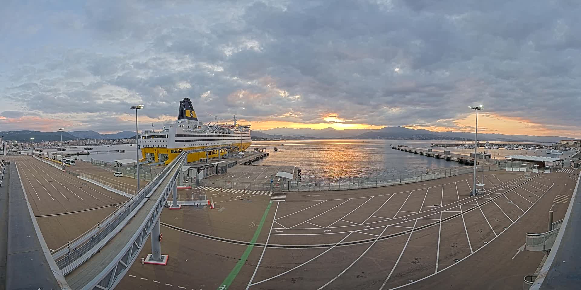 A large ferry is docked at a port during a sunset under a partly cloudy sky, with a walkway leading to the vessel and an empty parking lot nearby.
