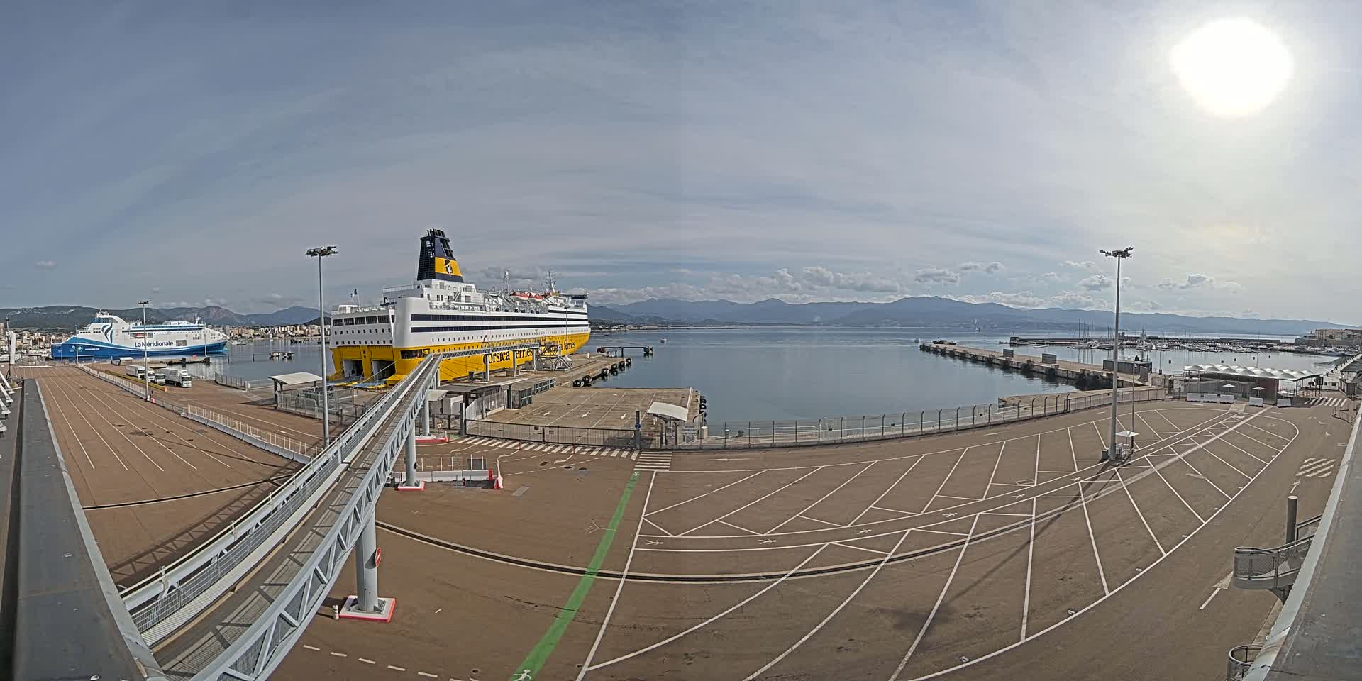 A large yellow and blue ferry is docked at a port on a sunny day, with another smaller ferry visible in the background, mountains in the distance, and a mostly empty parking lot in the foreground.