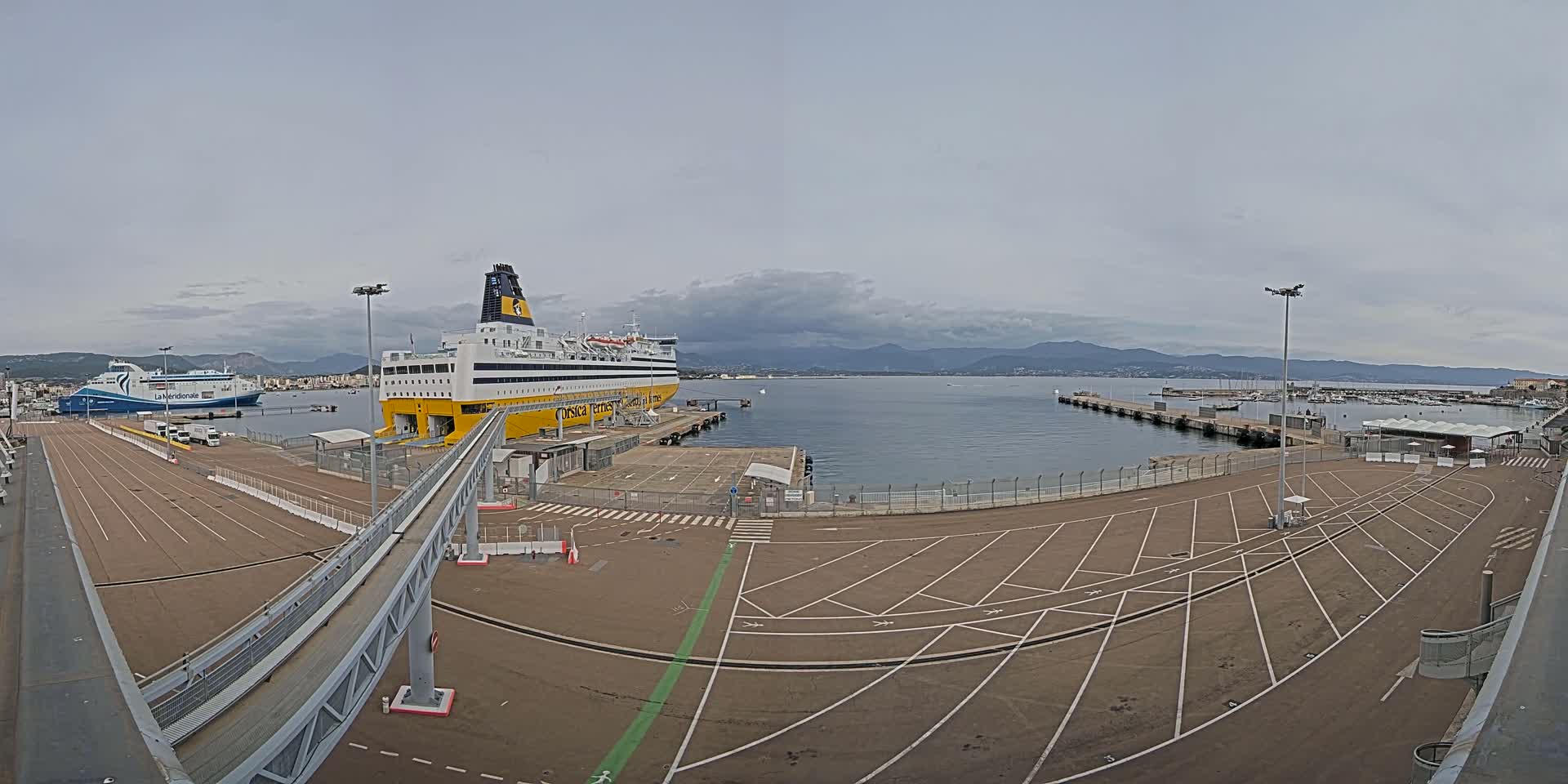 A large ferry is docked at a port on an overcast day, with another ferry visible in the background and a large empty parking lot in the foreground.