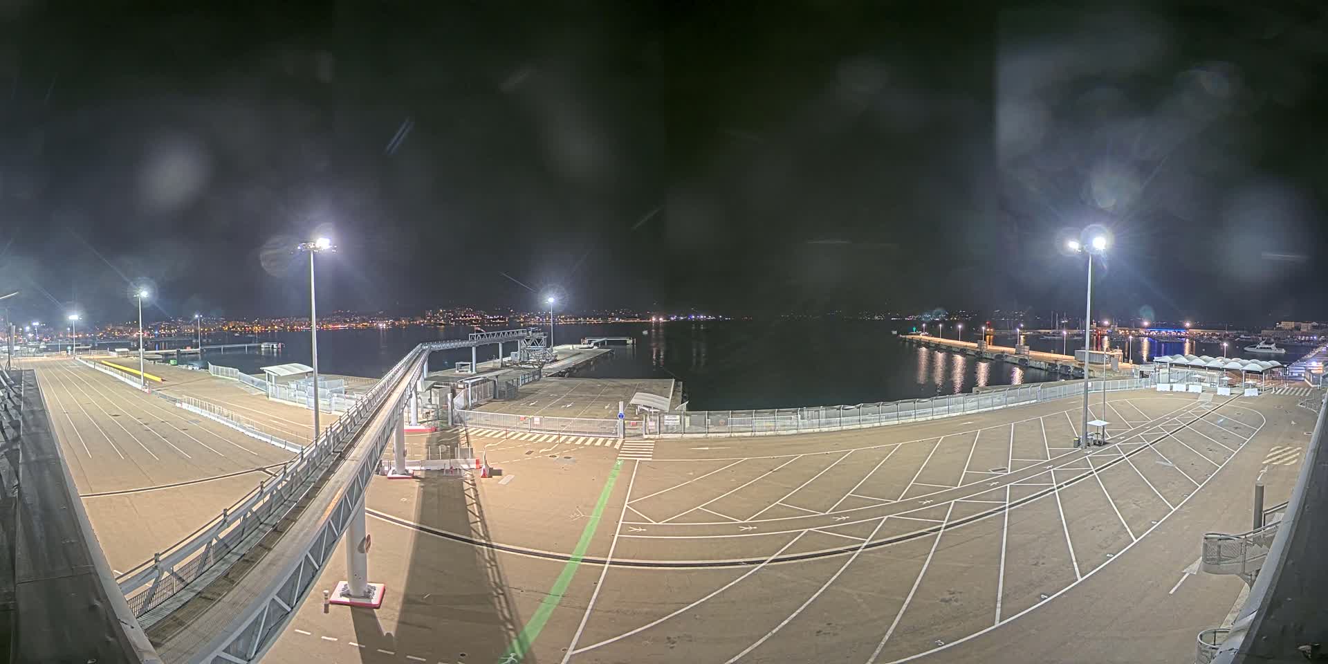 A nighttime view of an empty port area with a walkway and parking lot, illuminated by tall lights, overlooking a calm body of water and a distant city skyline.