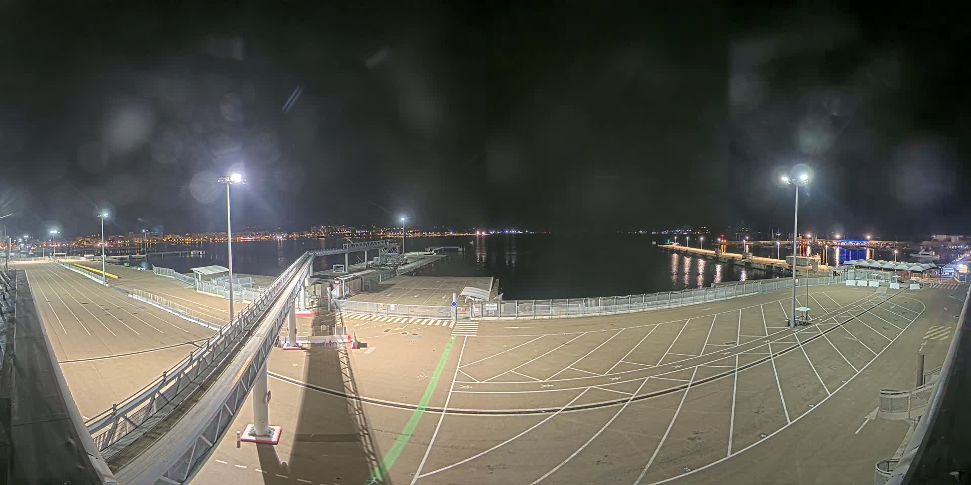 An empty, well-lit ferry terminal at night overlooks a calm body of water with a city skyline faintly visible in the distance under a dark, slightly overcast sky.