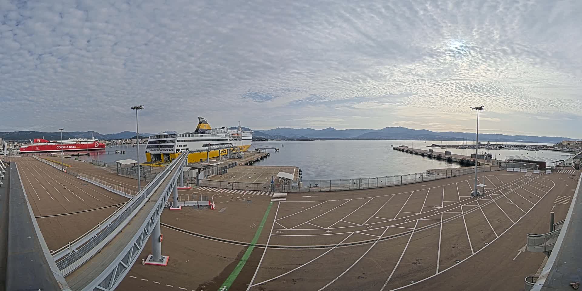 A ferry terminal with two ferries docked, an empty parking lot, and a calm body of water under a cloudy sky.