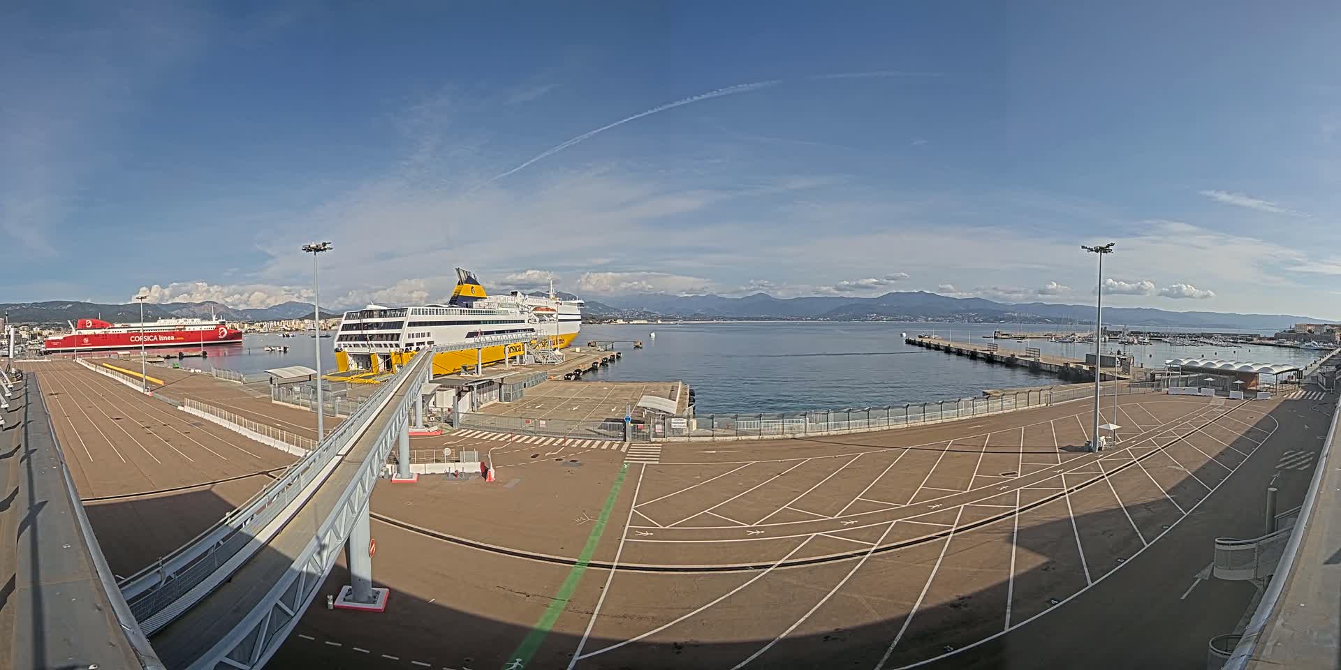 A mostly empty ferry terminal with two large passenger ferries docked, under a mostly clear sky with distant mountains visible across calm water.