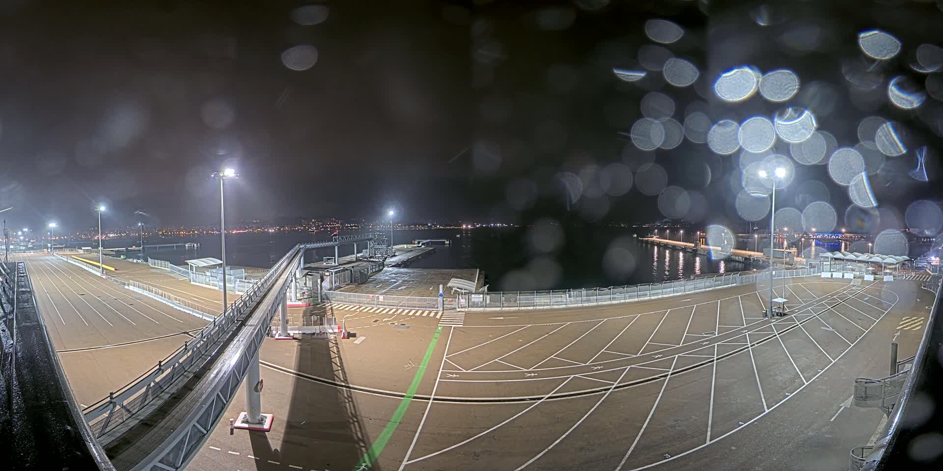 A nighttime view of an empty ferry terminal, illuminated by lights, with rain obscuring the view and a city skyline in the distance.